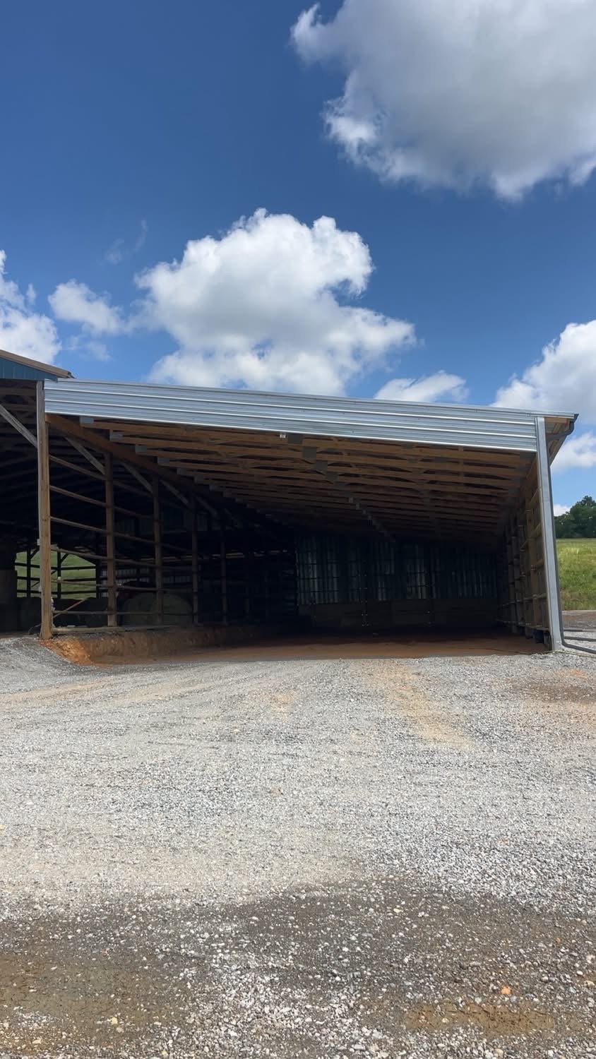 Open livestock barn with a metal roof, wooden frame, and gravel driveway under a cloudy sky.