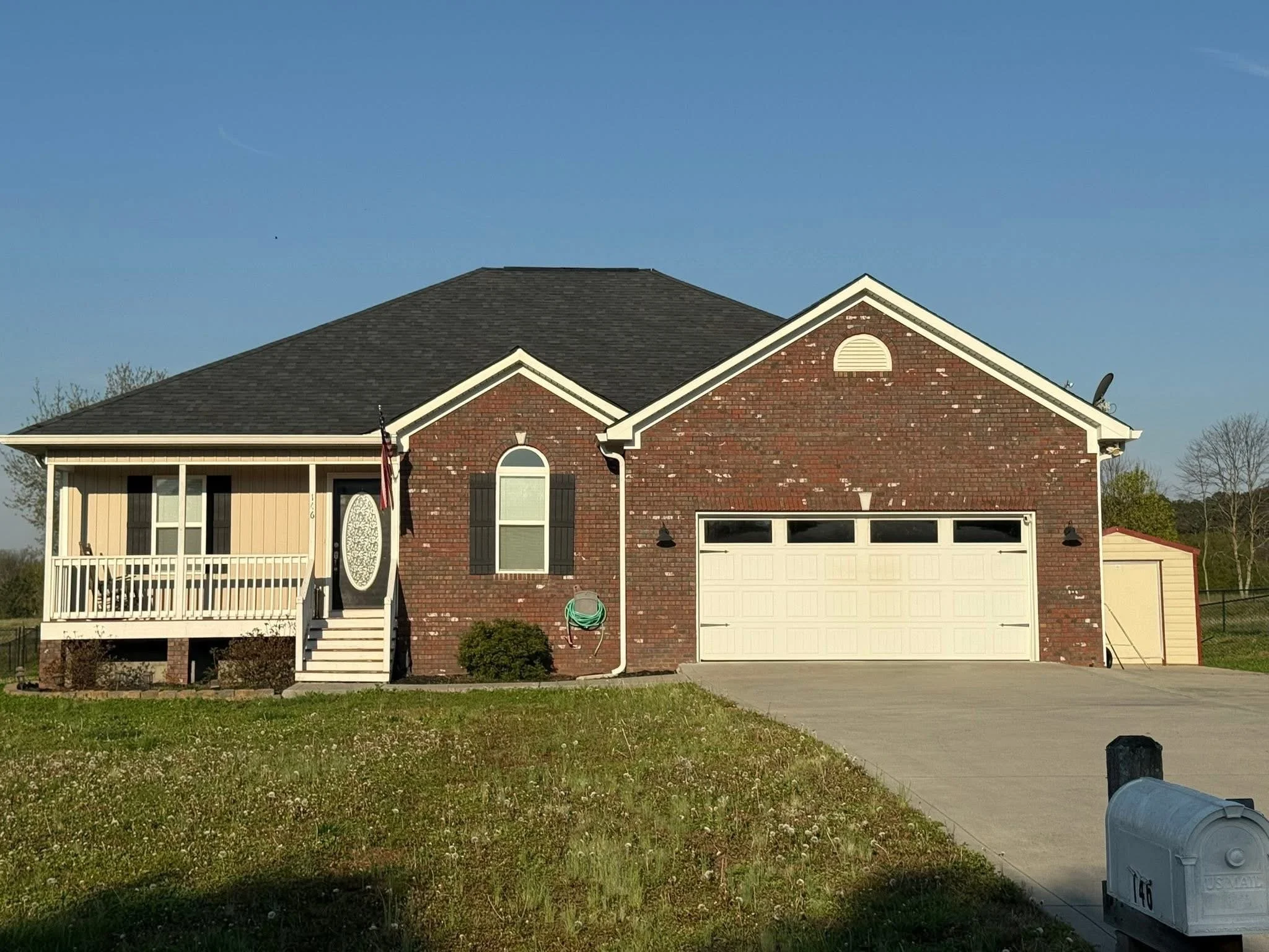 A two-story house with a brick facade and a garage, white porch railing, steps leading up to a front door, and a lawn with a garden hose, mailbox, and trash bin in front under a clear blue sky.