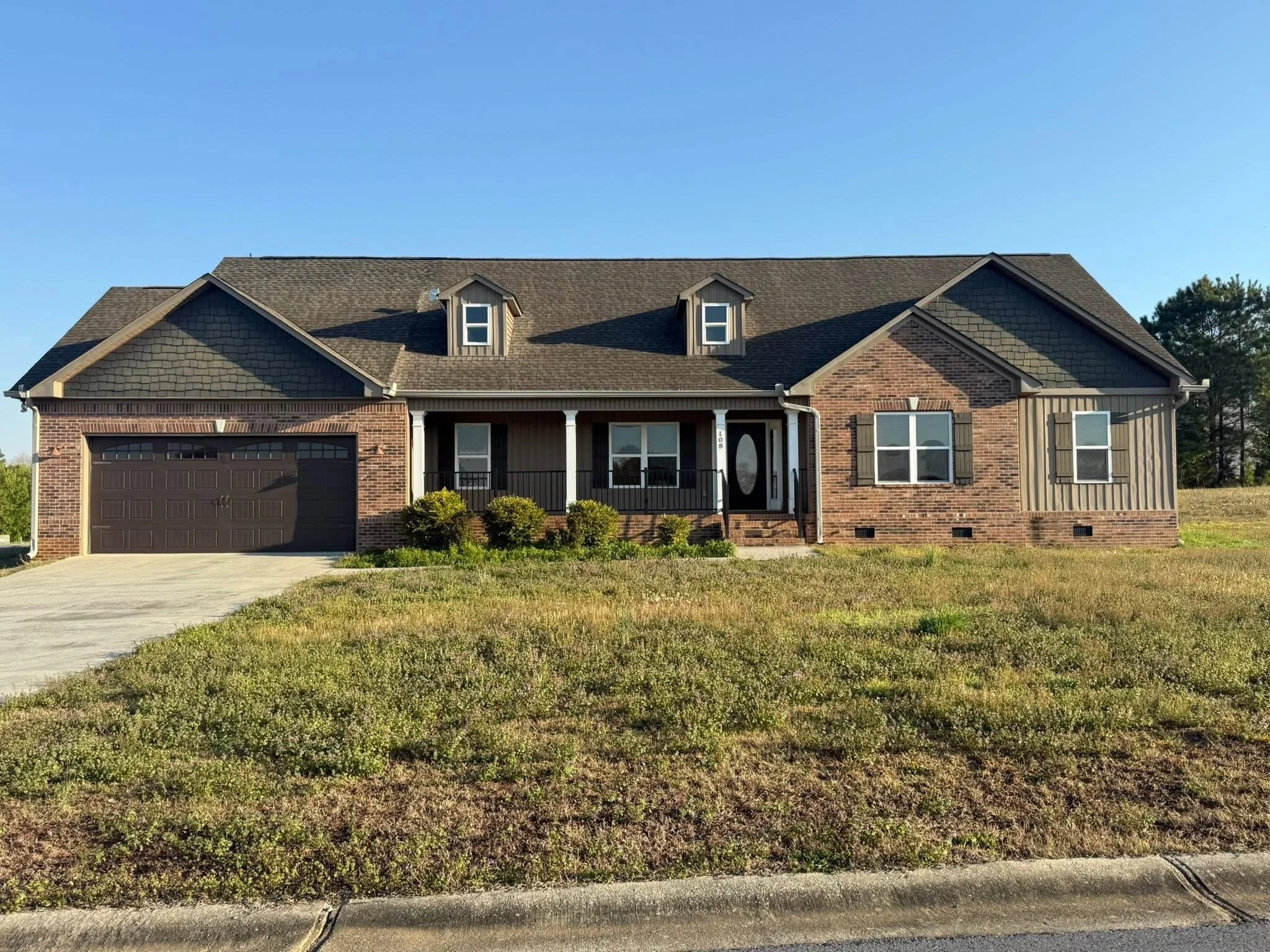 Front view of a modern brick house with a two-car garage, front porch, and dormer windows on a sunny day.