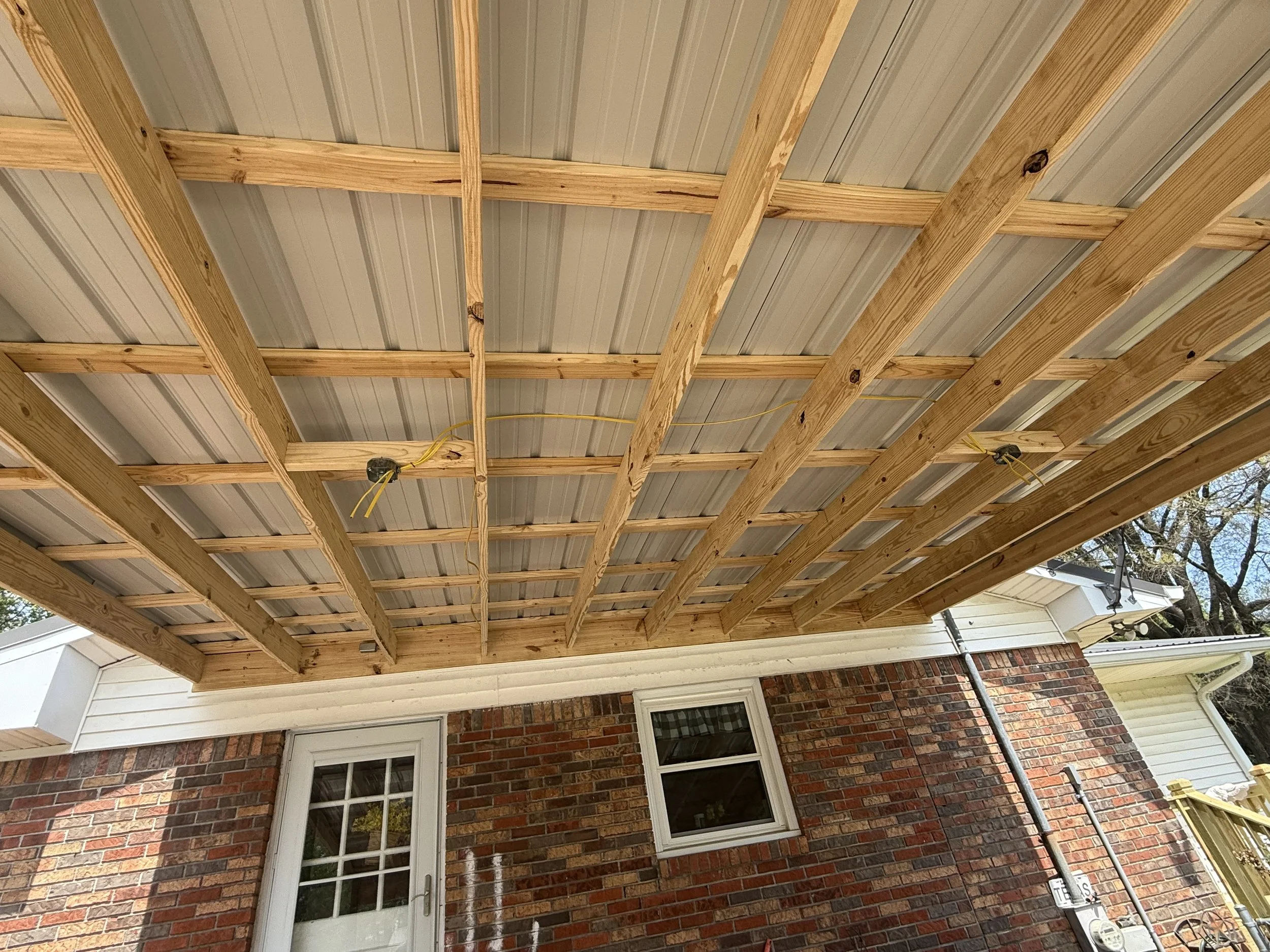 Construction of a wooden deck extension on the back of a brick house, with visible window and door, and trees in the background.