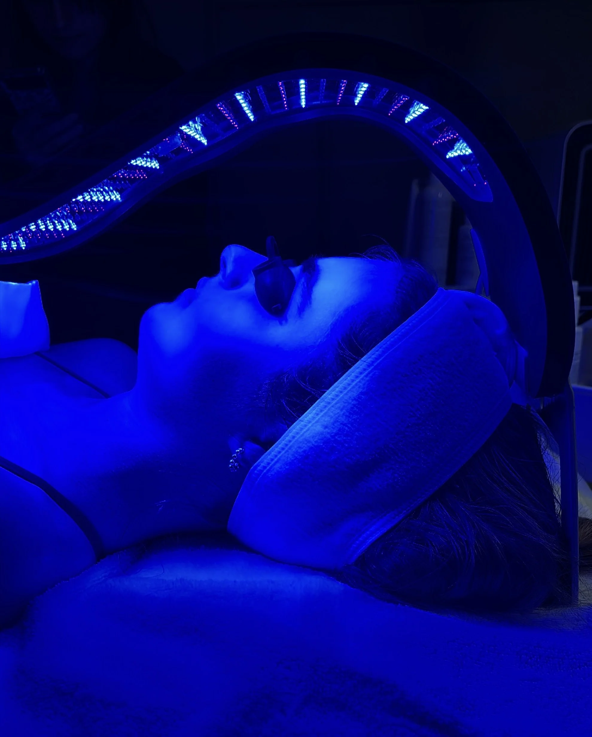 Person lying in a medical or aesthetic treatment bed, illuminated by blue LED lights, possibly during a beauty or skin treatment.