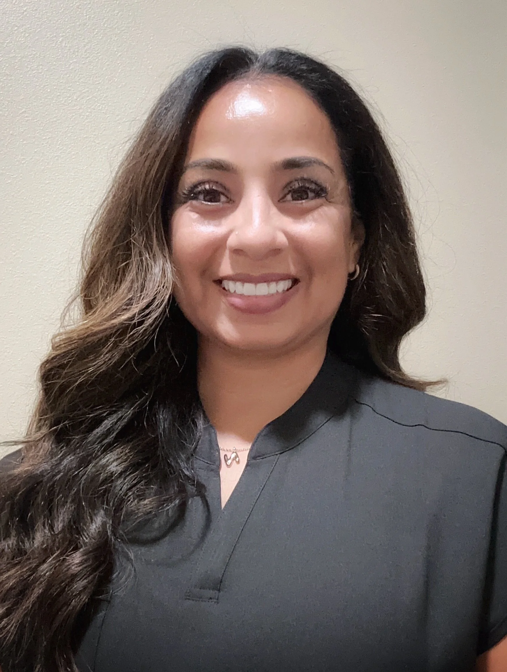 Close-up of a woman with long, dark, curly hair smiling, wearing a dark top, a necklace, and earrings, against a beige wall.