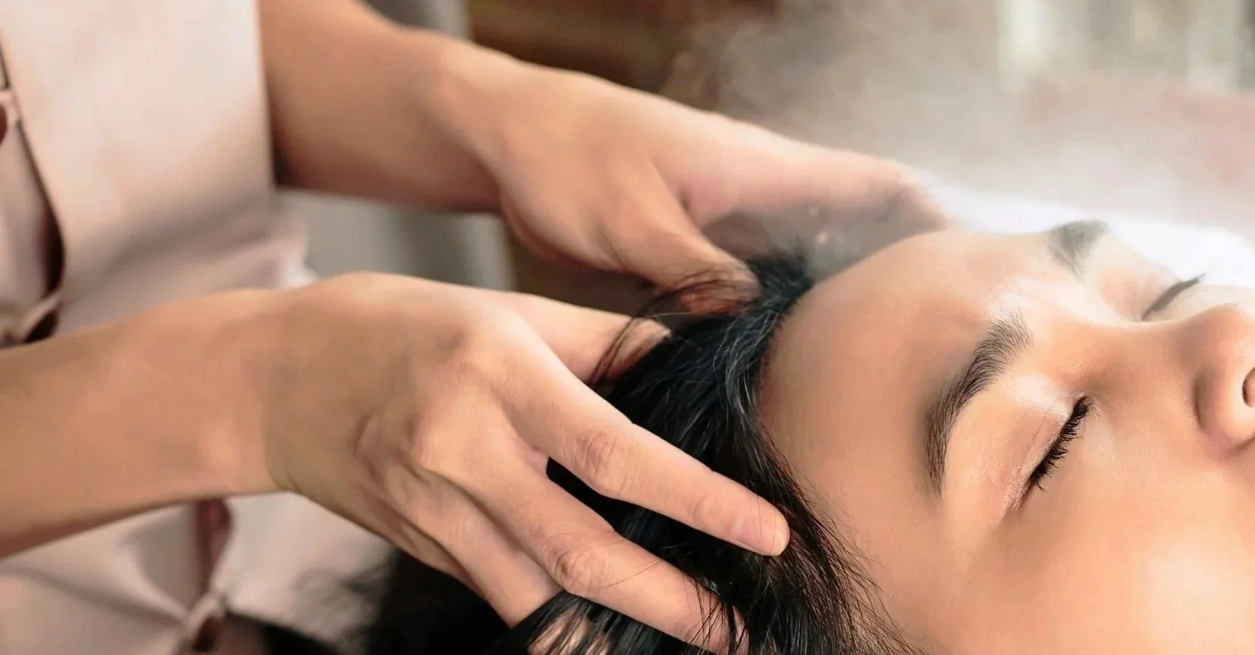 A person receiving a massage or scalp treatment, lying on their back with eyes closed, while another person gently massages their head with both hands.