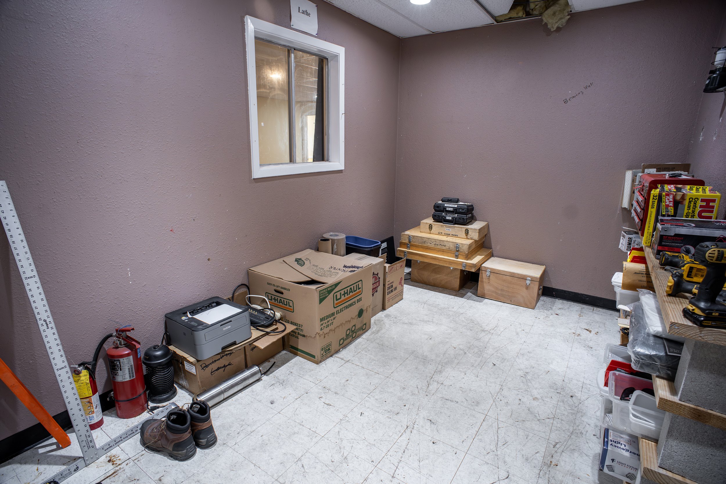 A cluttered room with pink walls, containing boxes, tools, shoes, and various supplies, including a small window and a fire extinguisher.