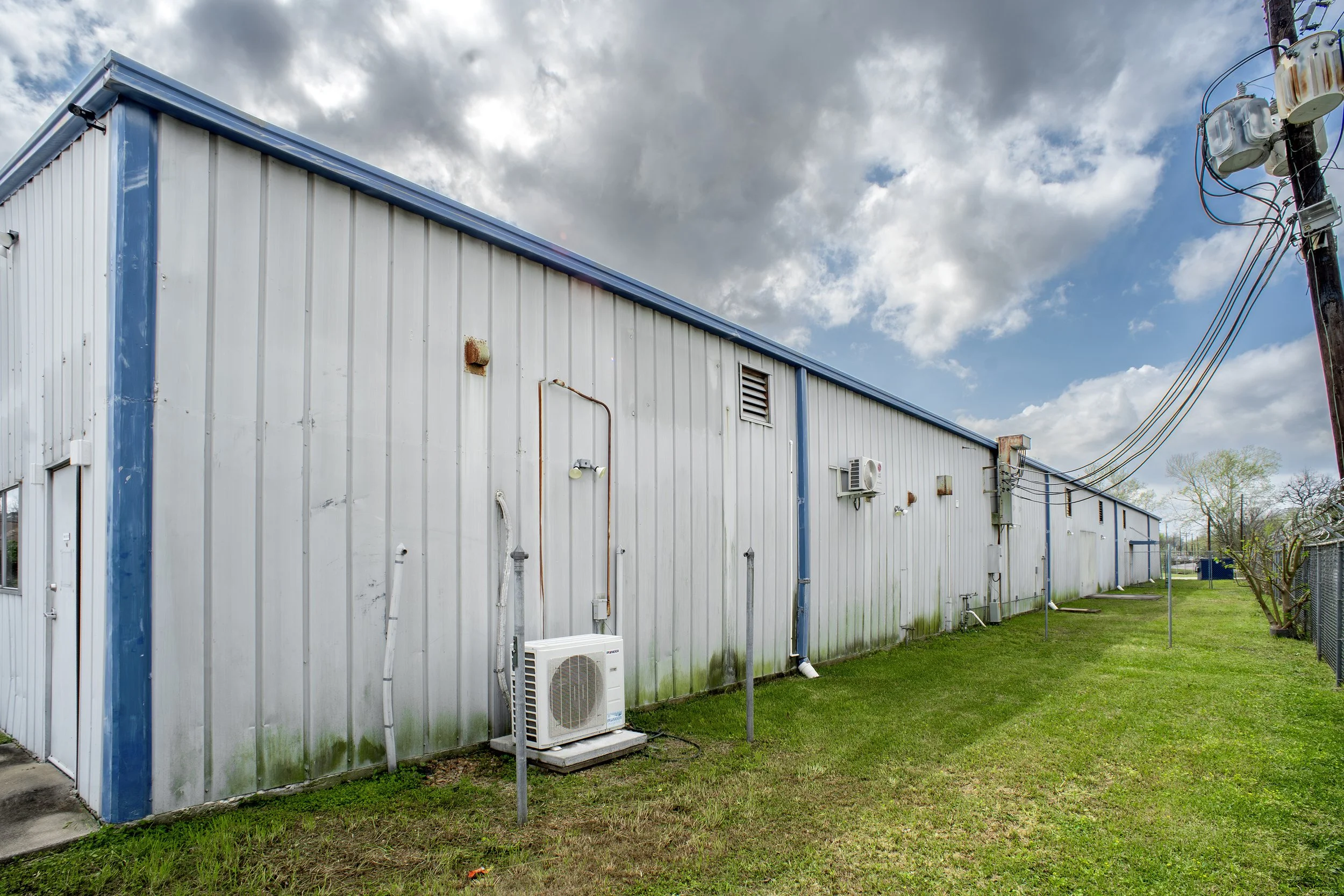 Side of a metal industrial building with HVAC unit, pipes, and vents, outdoors on a cloudy day, with grass and a fence.