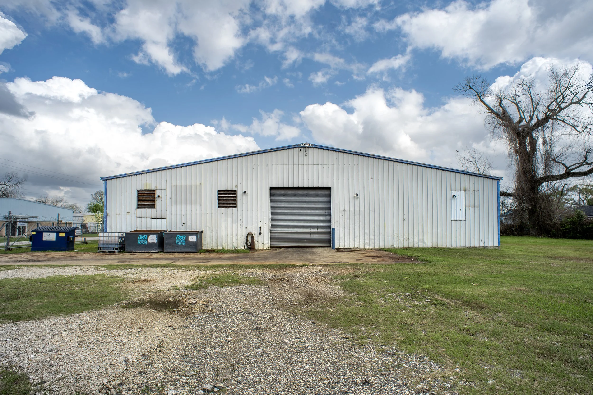 A white industrial building with a blue roof and rolled-up garage door, set in a grassy area with a gravel driveway, tree on the right, and partly cloudy sky overhead.