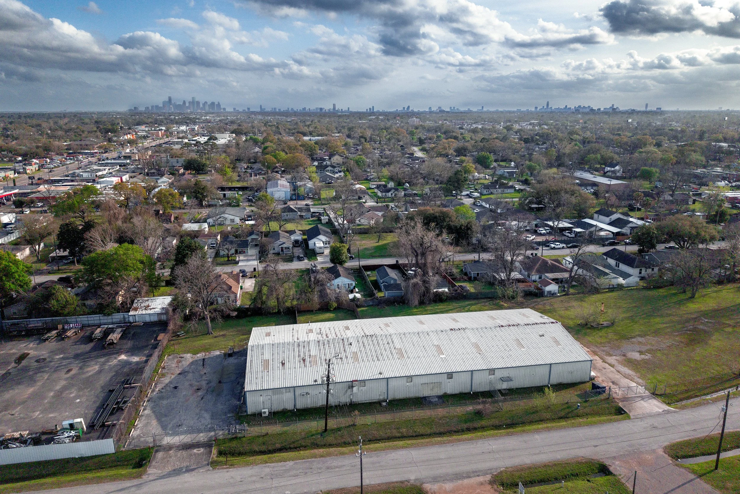 Aerial view of a suburban area with houses and trees, with a city skyline visible in the distance under cloudy skies.