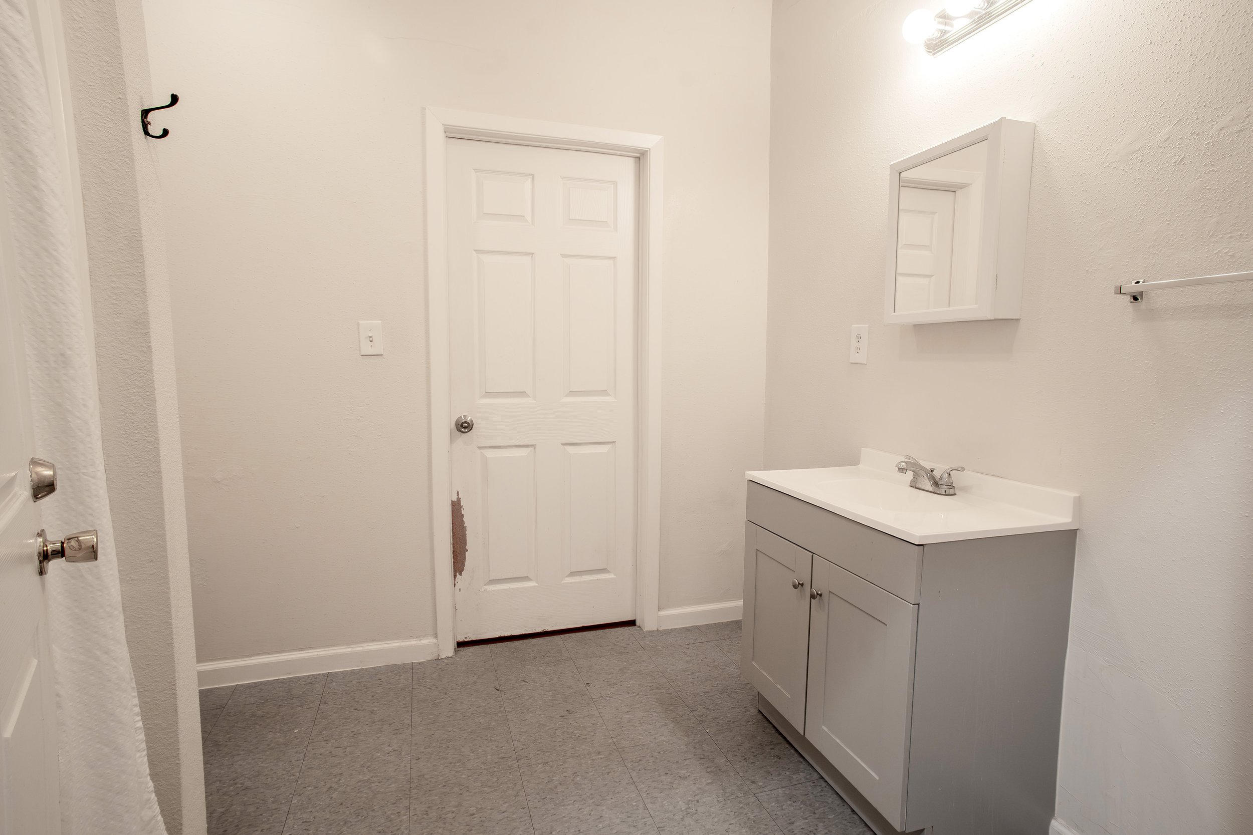 A small bathroom with a white door, a white vanity with a sink, a mirror, and a wall-mounted light fixture. The bathroom has beige textured walls and a gray tile floor.