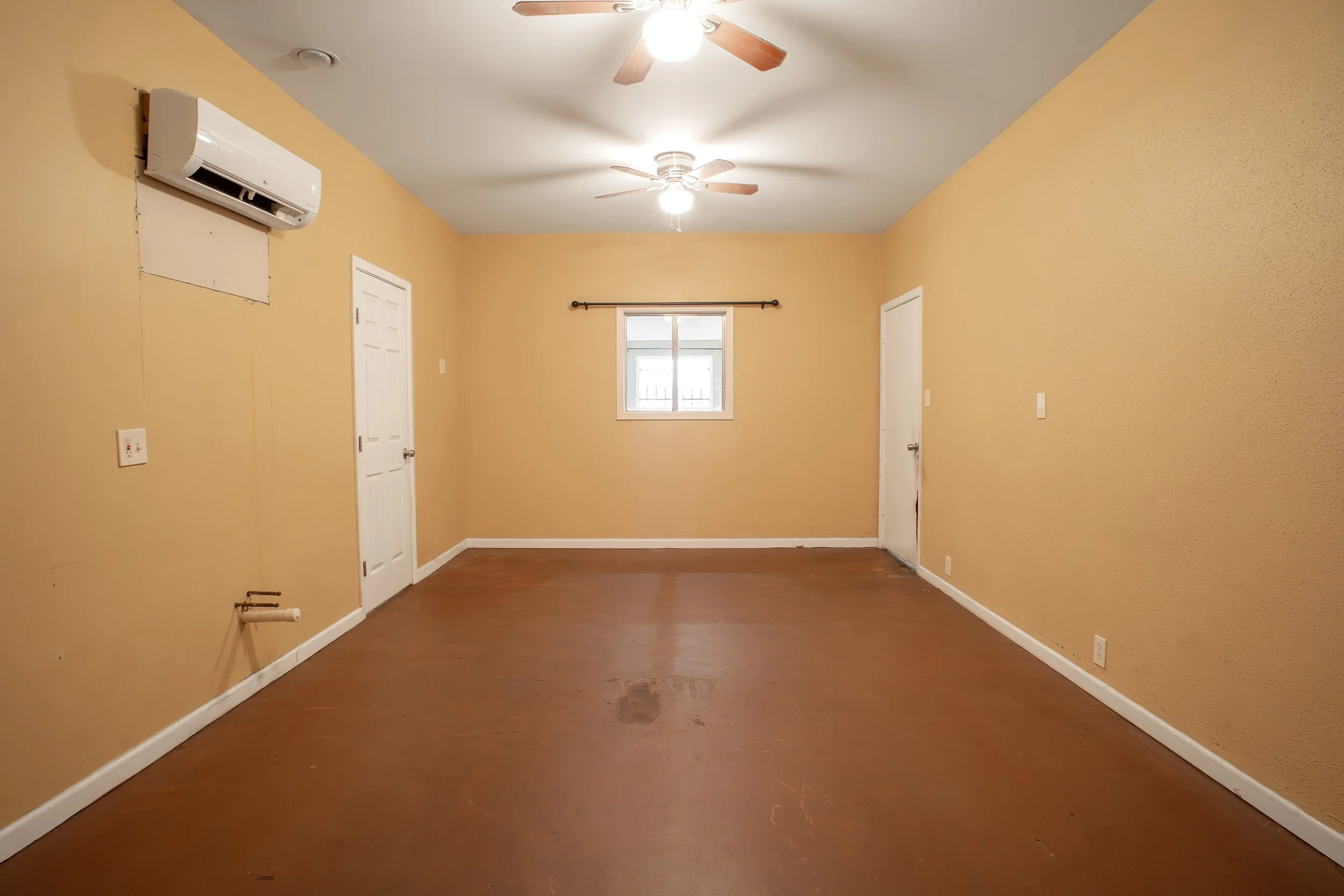 Empty room with yellow walls and hardwood floors. There is a window with a black curtain rod, a ceiling fan with wooden blades, and an air conditioning unit on the wall with a patch of drywall below it. There are two closed doors, and electrical outl