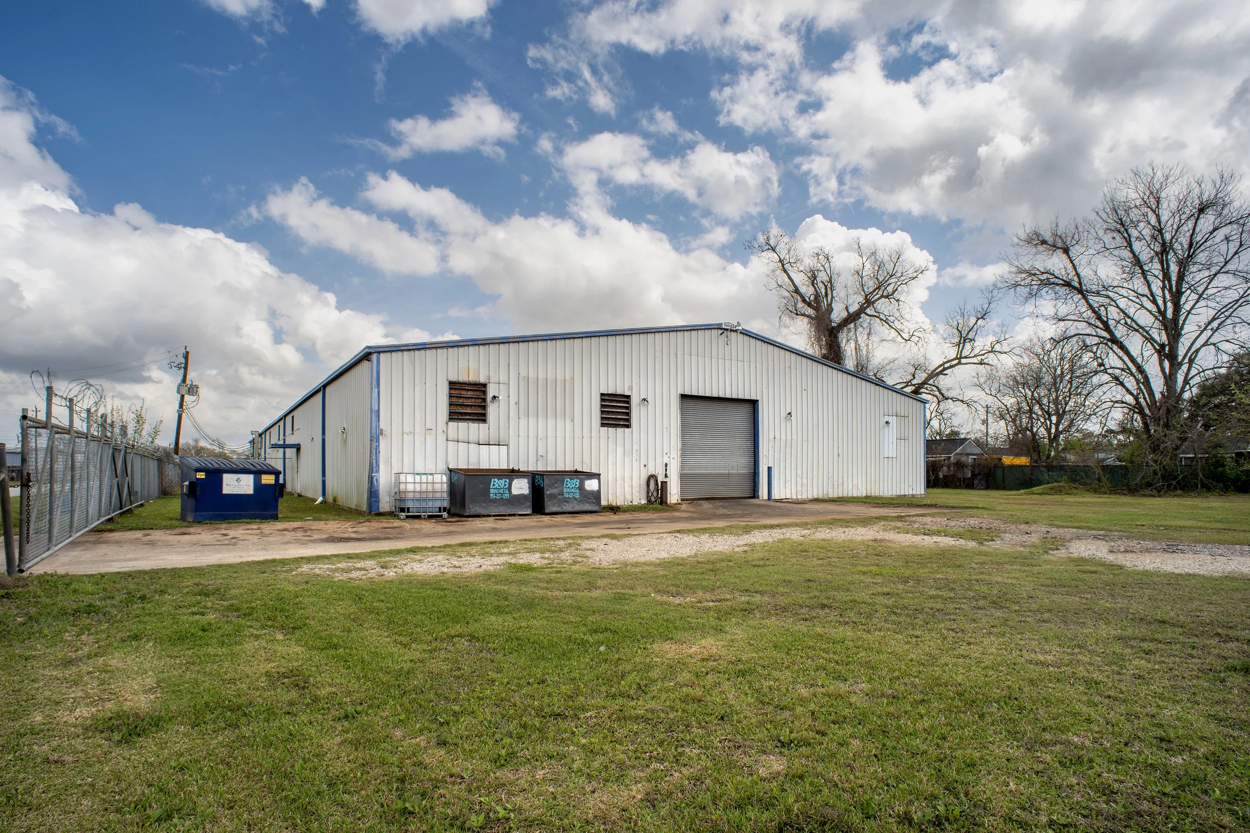 A metal building with a garage door, vents, and a couple of trash bins in front, set against a partly cloudy sky with some tall, leafless trees and a grassy area.