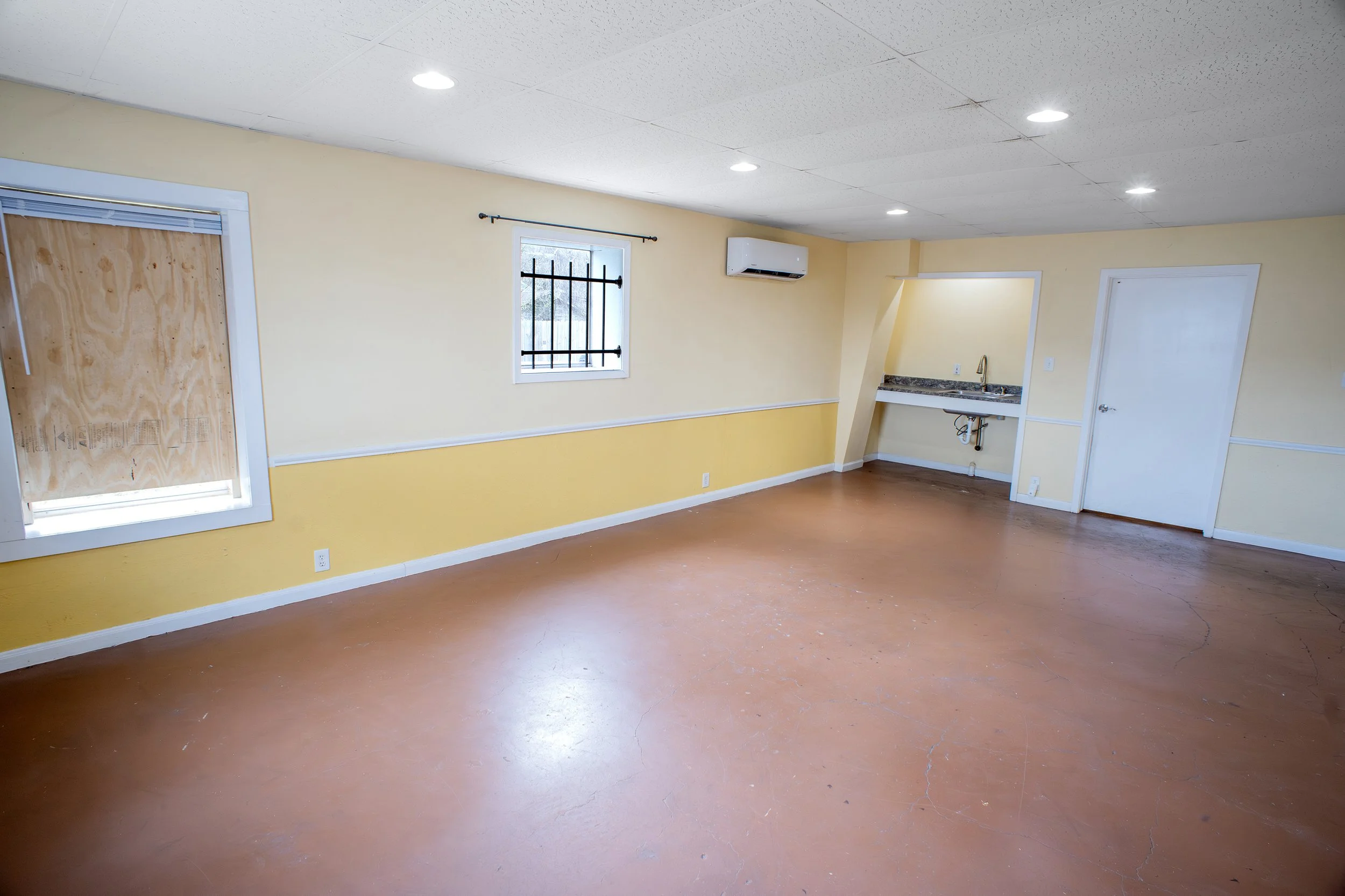Empty room with yellow walls, brown floor, window with security bars, and a small sink area with exposed plumbing.