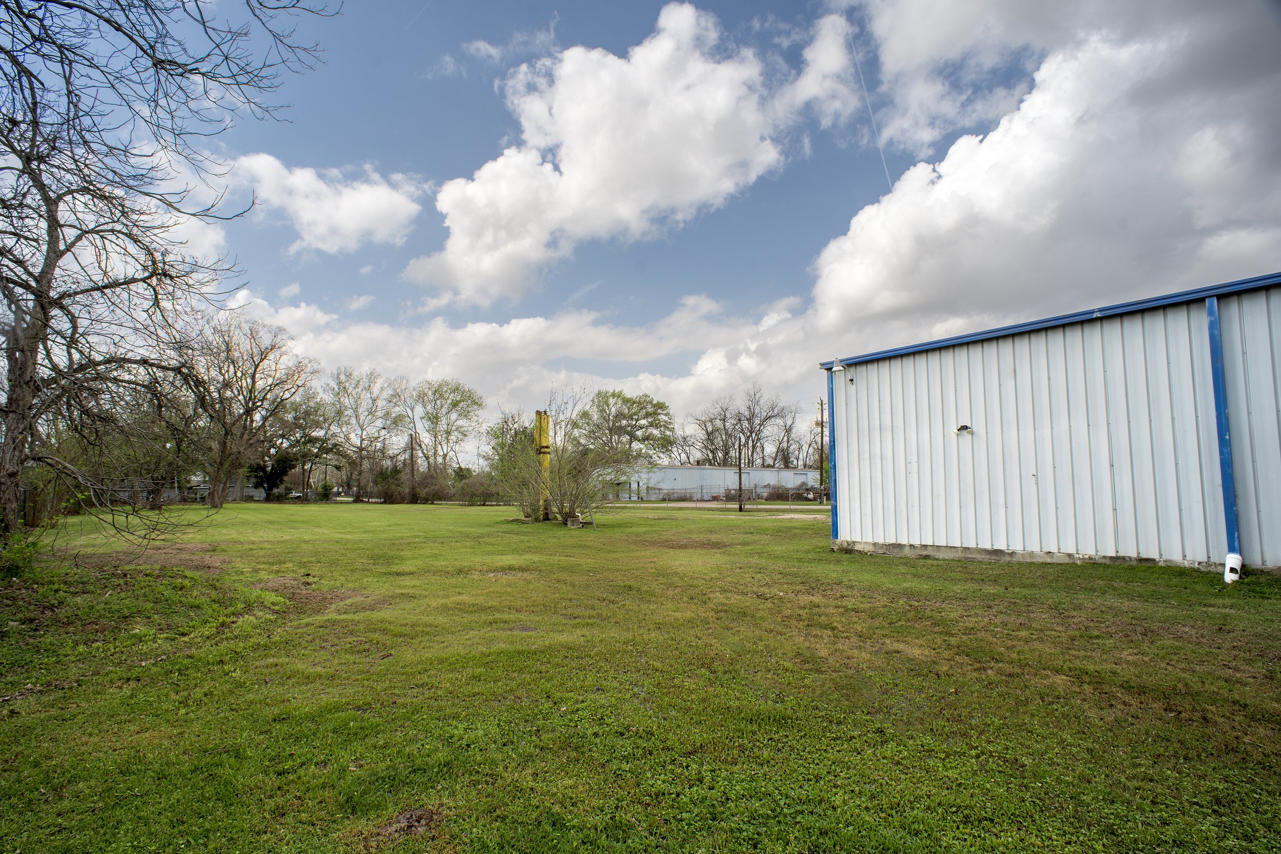 A grassy field with trees, a building with a blue roof on the right, and a partly cloudy sky overhead.