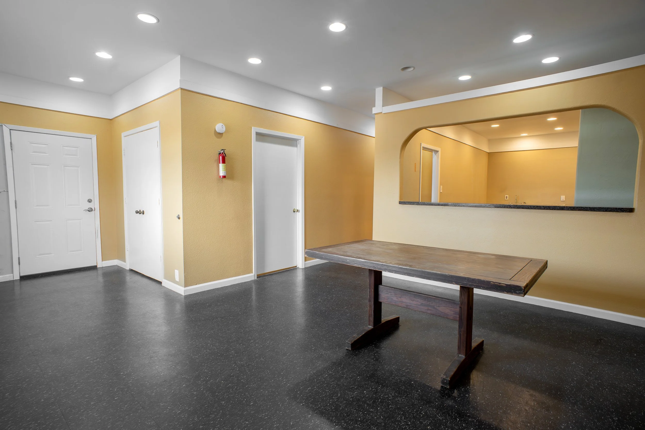 Interior of an apartment or office space featuring white and yellow walls, a doorway, a fire extinguisher, ceiling lights, a granite counter window, and a wooden table on a dark, speckled floor.