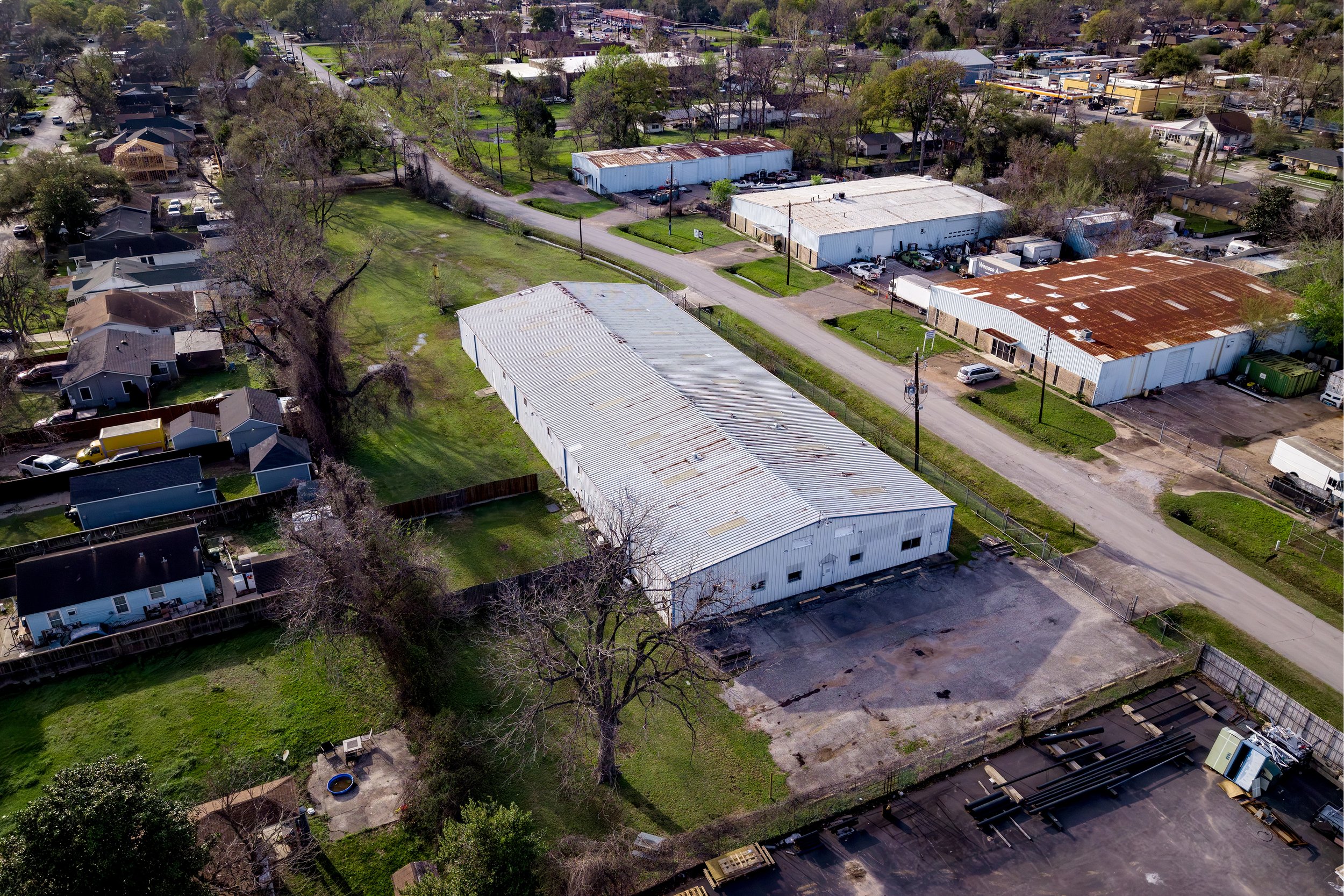 Aerial view of a semi-urban area with residential houses, commercial buildings with rusted roofs, and trees with some spring foliage. Streets run through the neighborhood, with cars parked along the roads and in parking lots.