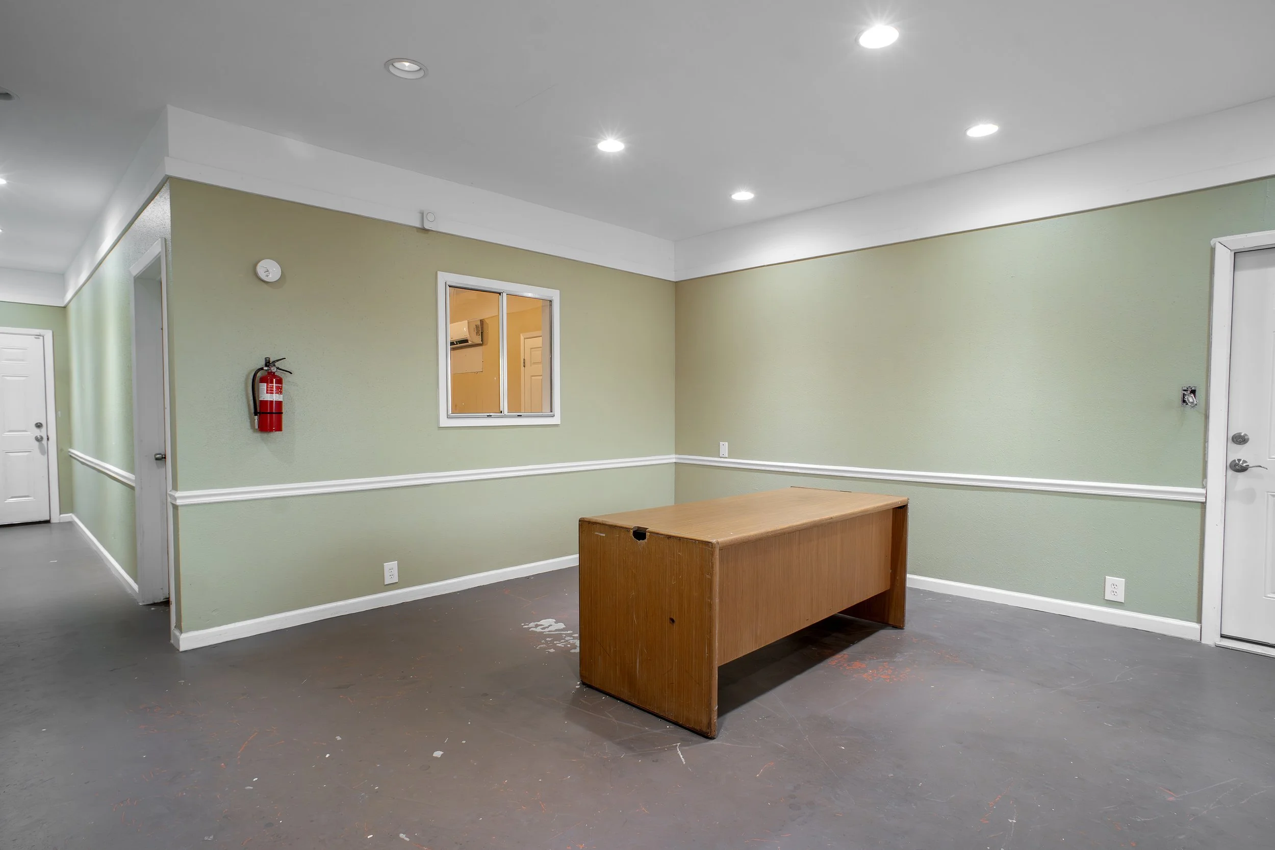 Empty room with green walls, round ceiling lights, a small wooden table, a window, a fire extinguisher on the wall, and a door.