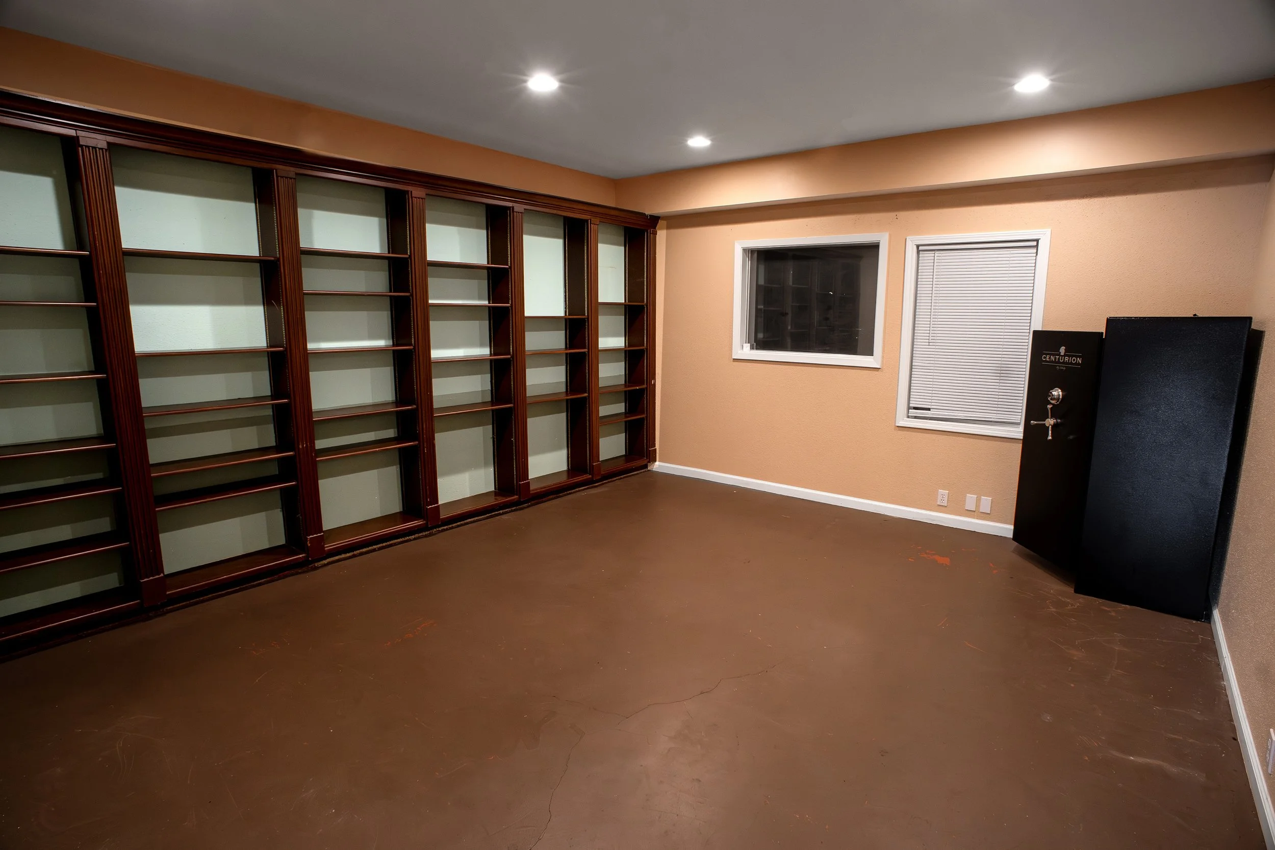 Empty room with large wooden bookshelf, window, and door with blinds, with a black safe resting against the wall.