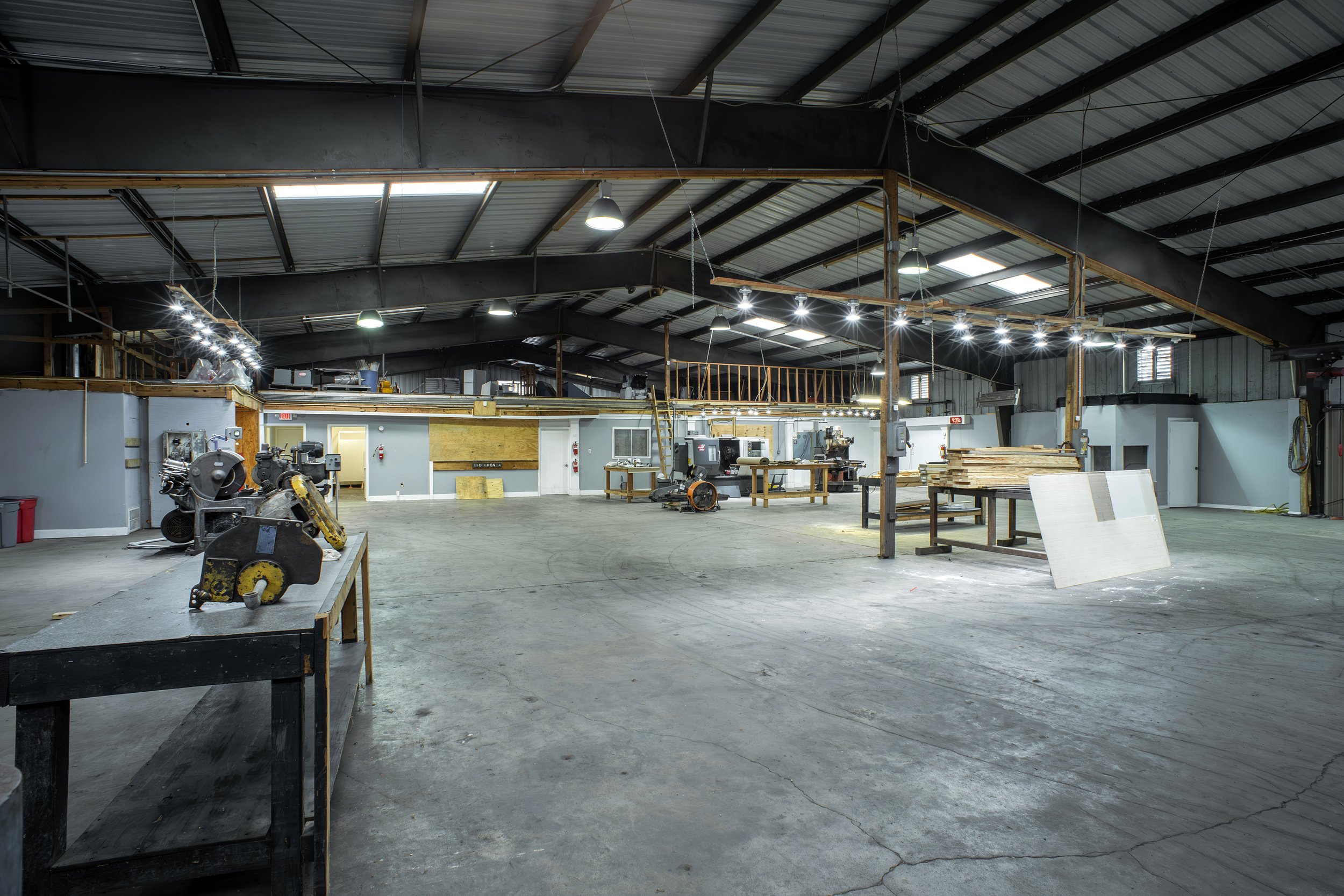 Interior of a woodworking or manufacturing workshop with machinery, workbenches, and wooden materials, inside a large warehouse with a metal roof.