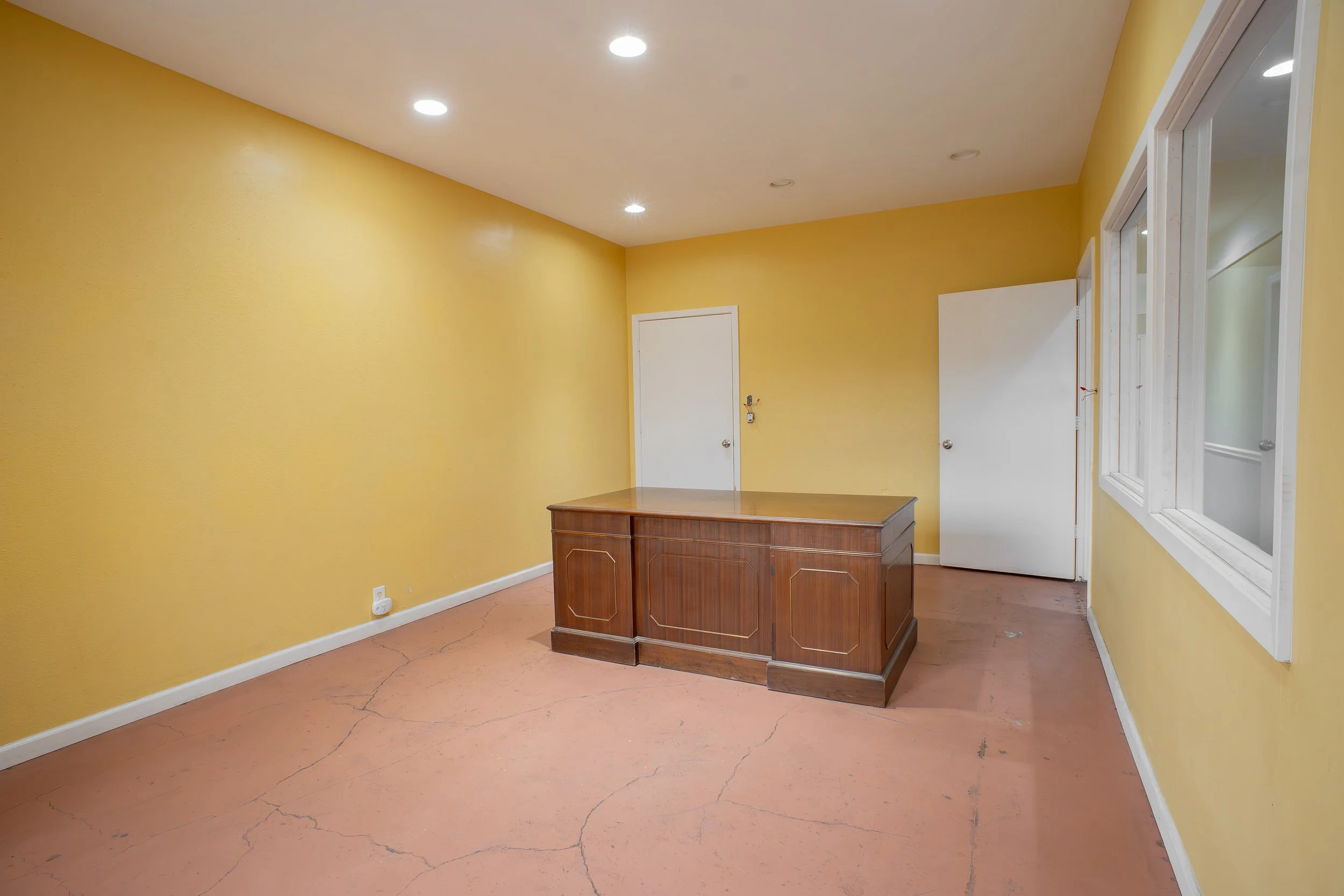 Empty room with yellow walls, a wooden desk, white doors, and windows, with a concrete floor and ceiling lights.