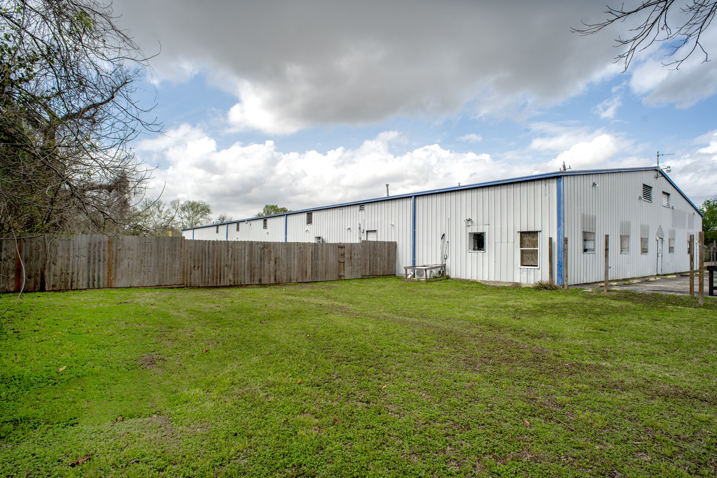 A large metal building with a blue roof surrounded by a wooden fence, with a grassy yard and leafless trees on the sides, under a partly cloudy sky.