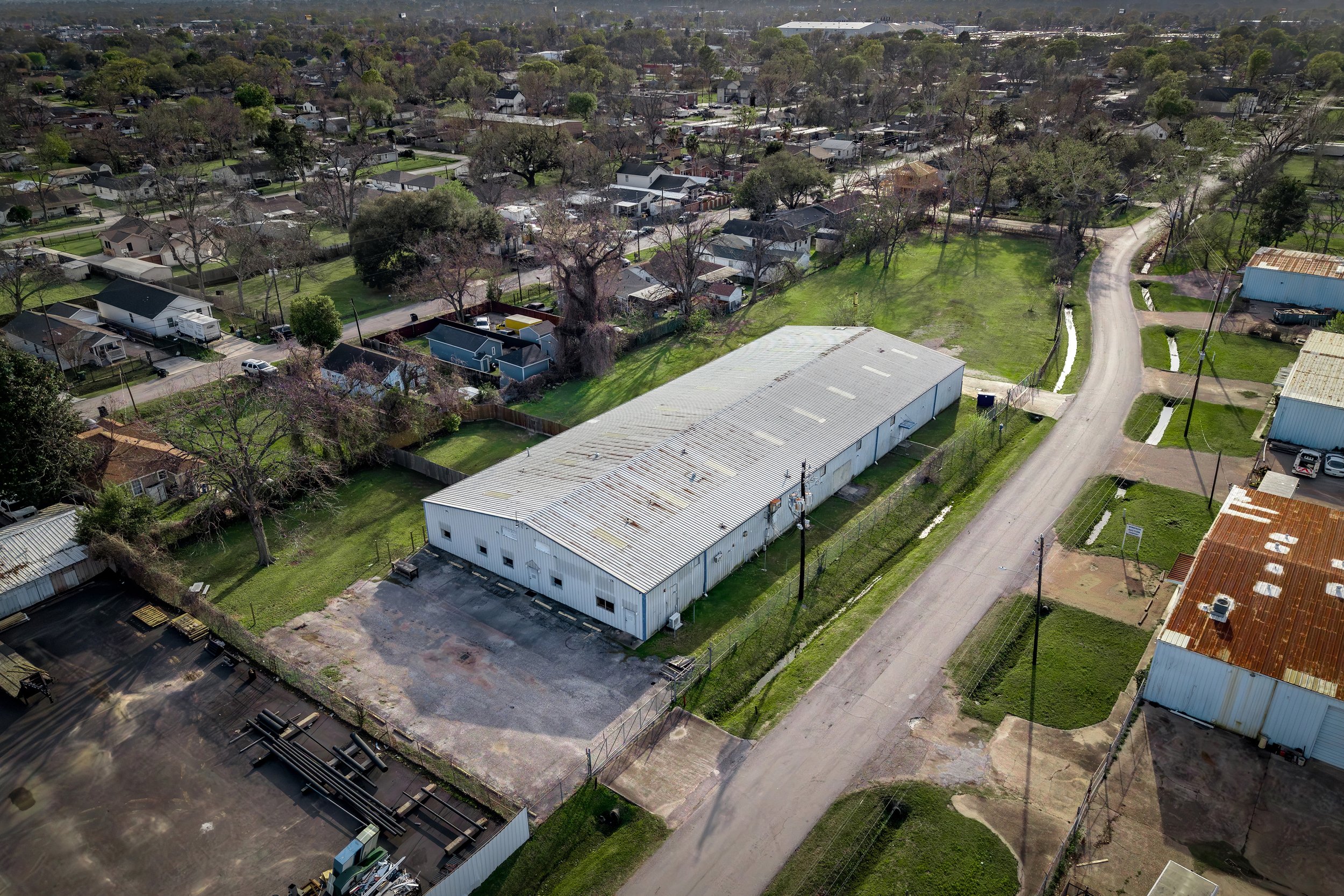 An aerial view of a neighborhood with houses, trees, and streets, including a large storage or industrial building with a metal roof in the foreground.