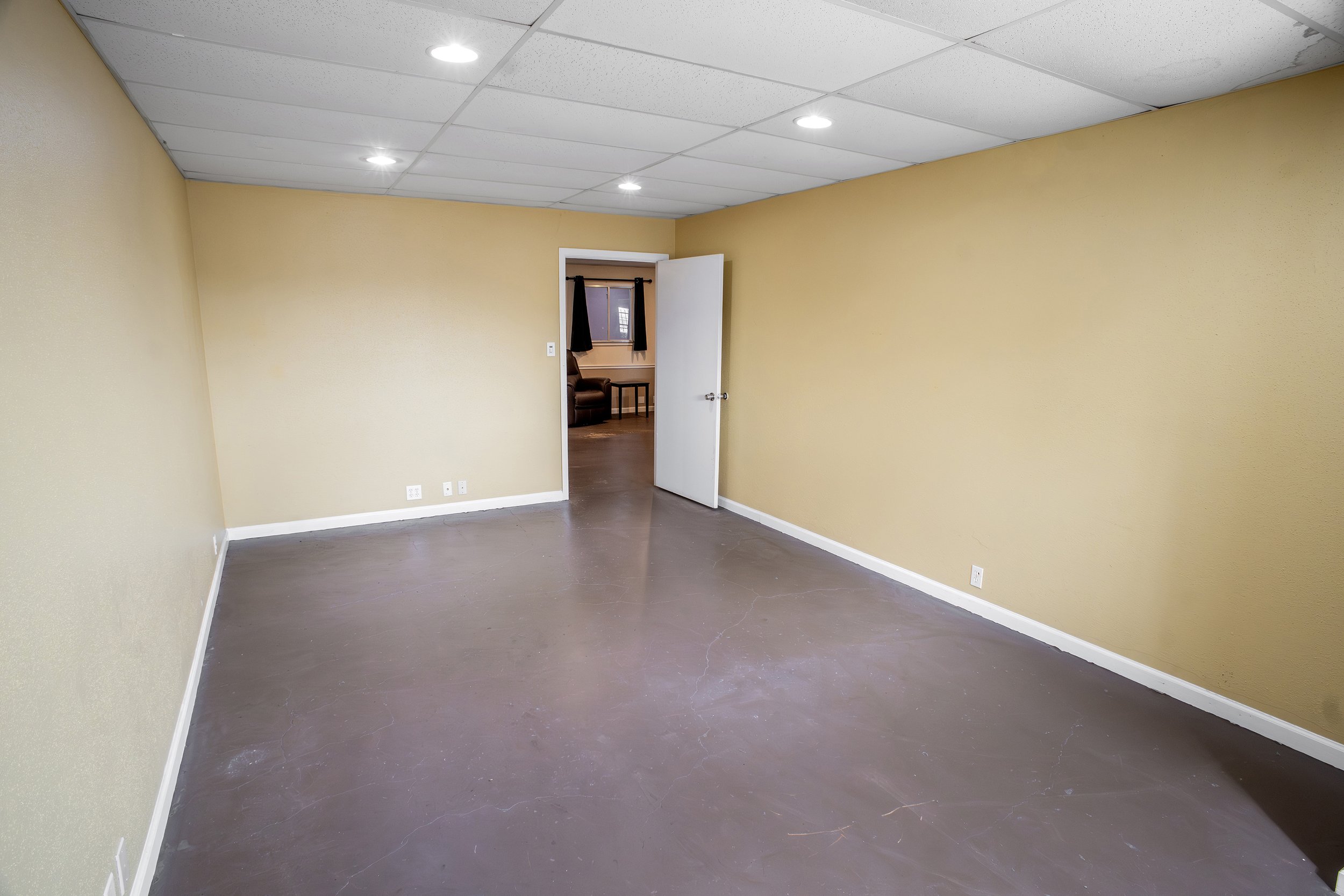 Empty room with beige walls, white ceiling tiles, and dark gray flooring. There is an open door leading to another room with a window, a brown chair, and a small table.