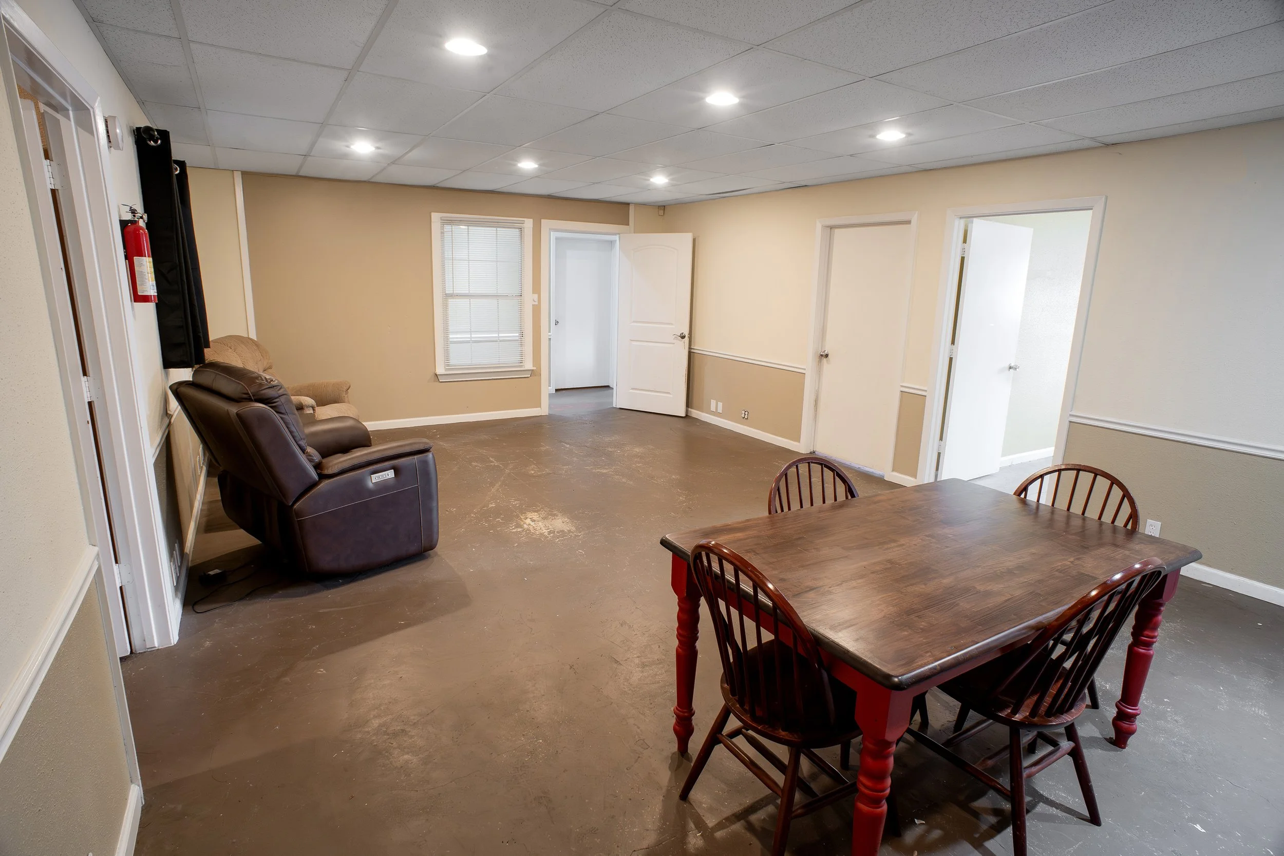 Interior of a room with beige walls, white ceiling tiles, wooden furniture, leather armchair, and several closed doors, with a fire extinguisher mounted on the wall.