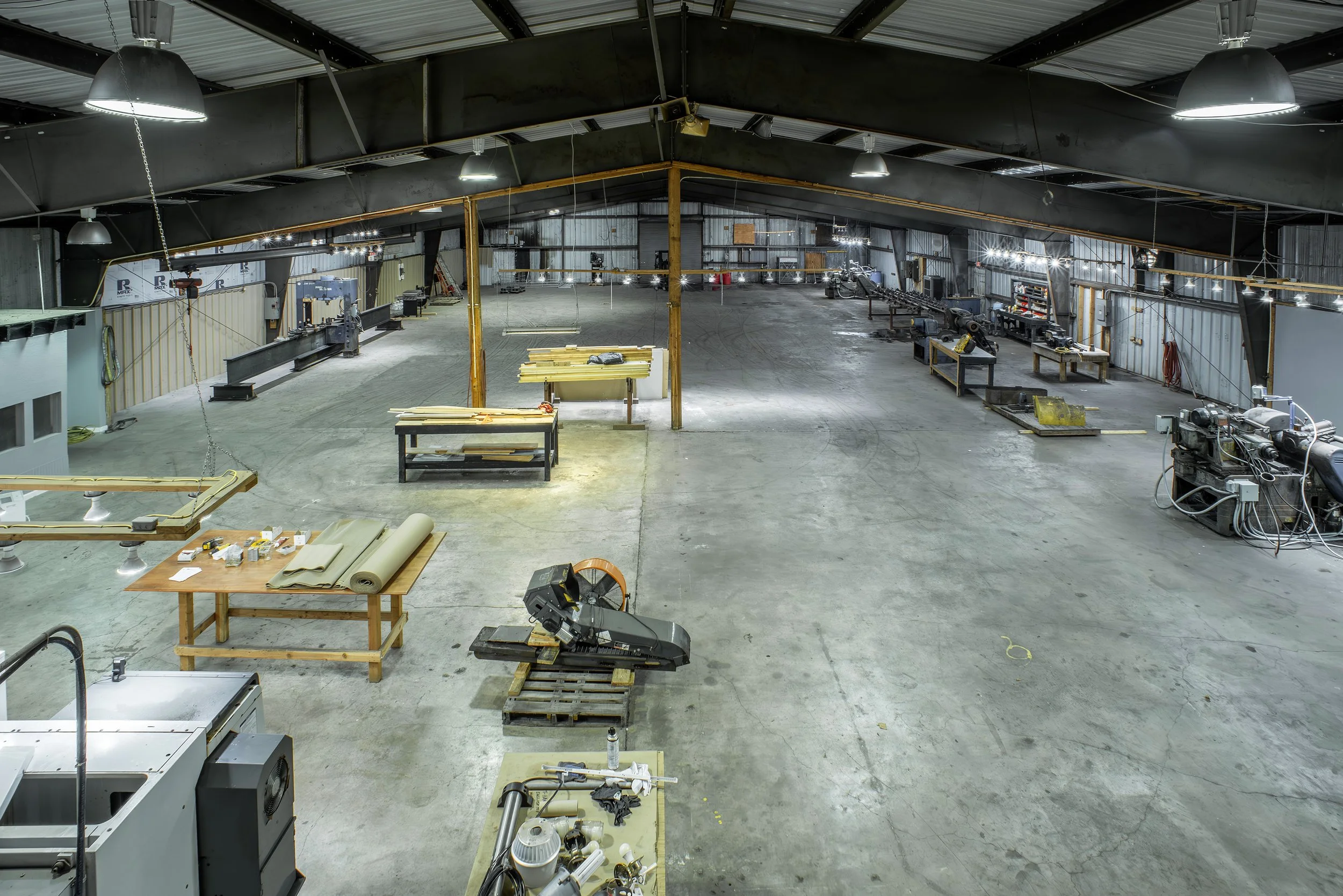 Interior of a large, empty industrial workshop or warehouse with metal walls and ceiling, various tools, equipment, and materials scattered on tables and the floor, and overhead lighting.