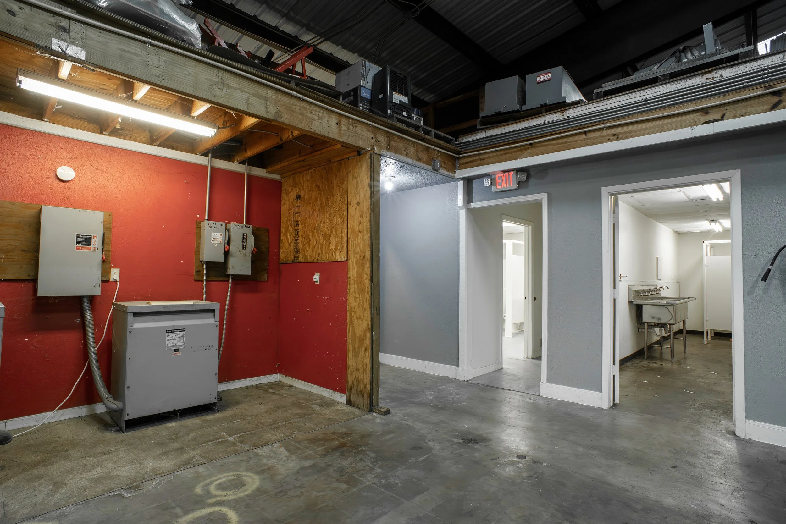 An industrial interior with a red wall, electrical panels, and a doorway leading to a room with a sink. Ceiling has exposed pipes and HVAC units.