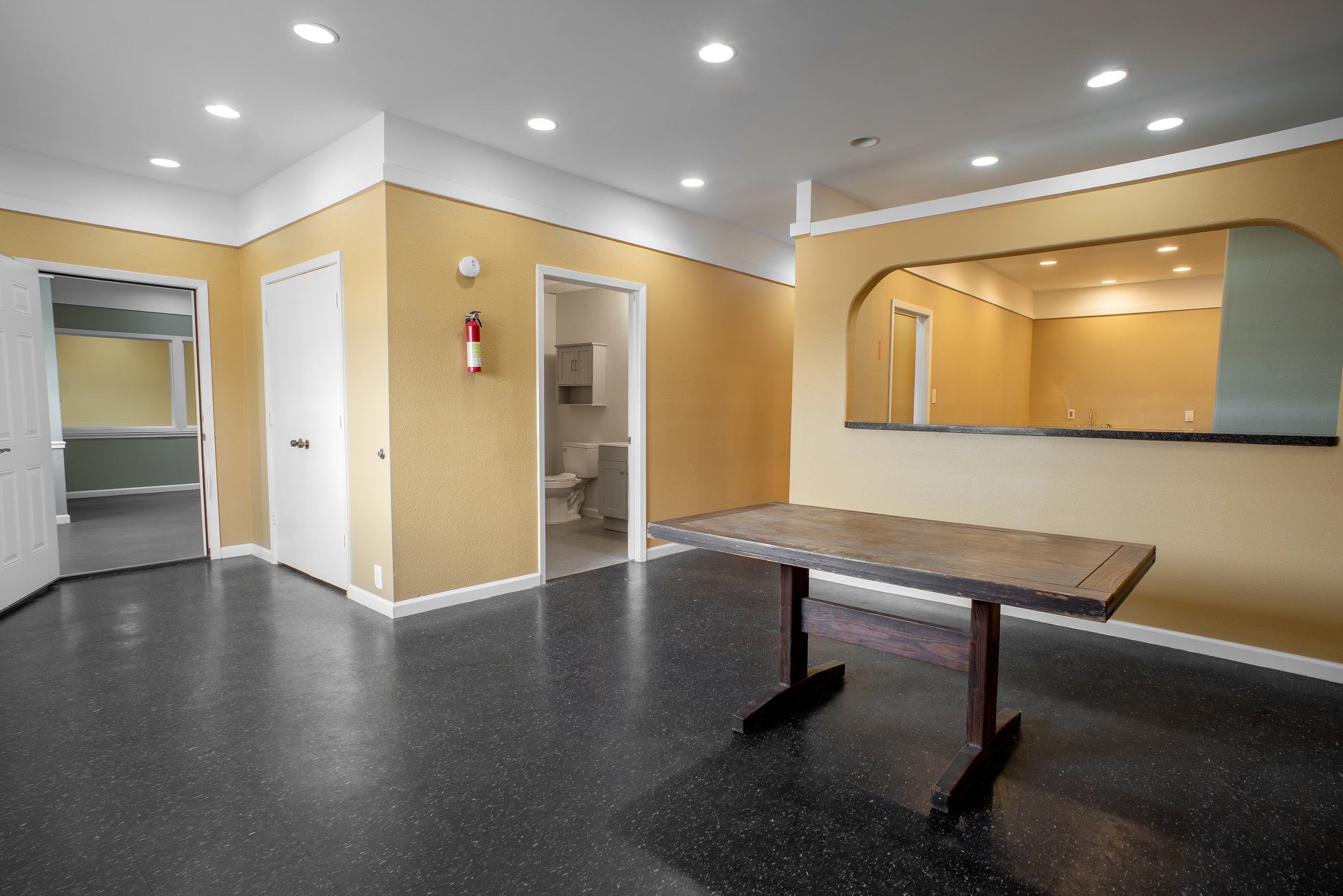 Interior of a room with yellow walls, ceiling with recessed lights, a wooden table, and doorways leading to other rooms, including a bathroom with a toilet and cabinet. Fire extinguisher mounted on wall.