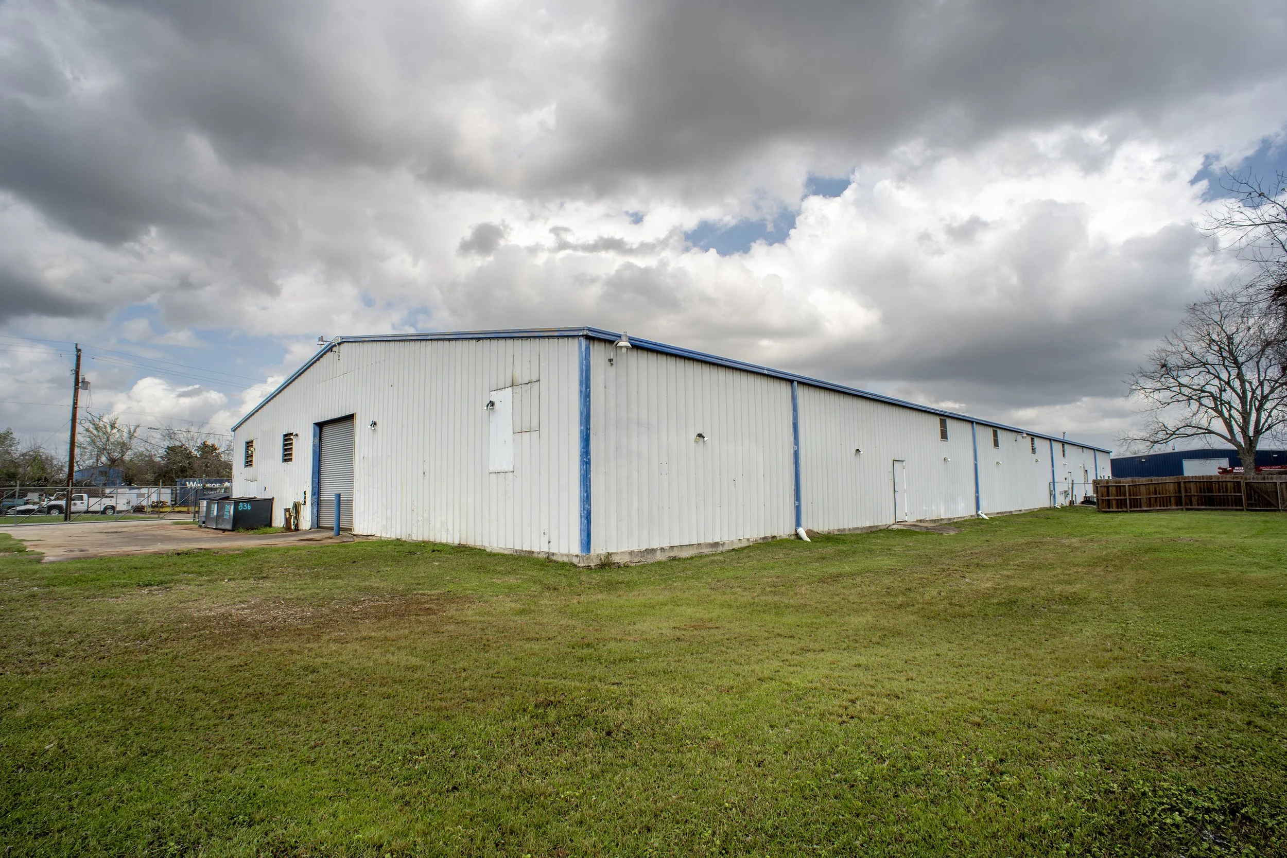 A large white industrial building with blue trim under a cloudy sky, surrounded by a grassy lawn.