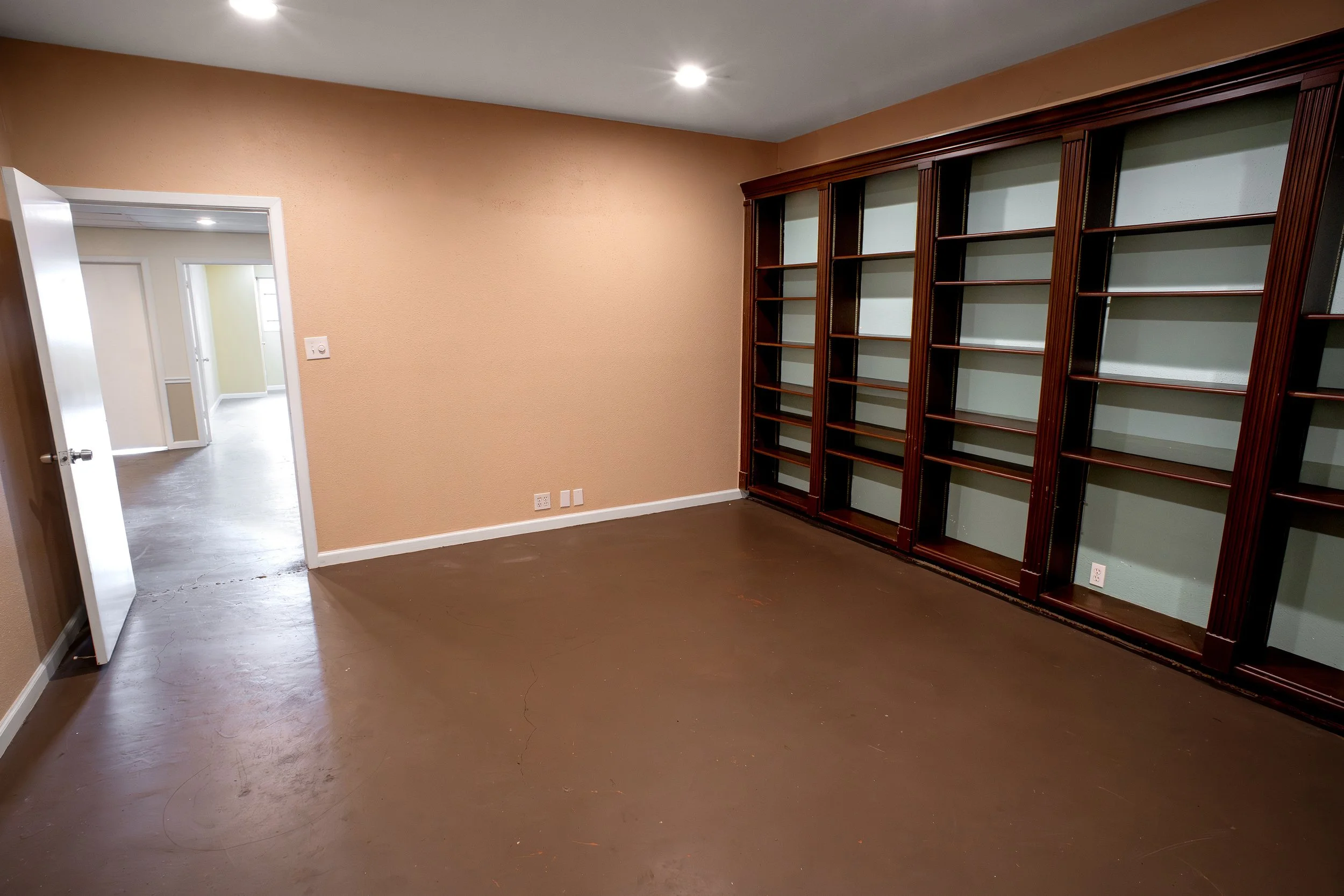 Empty room with a built-in wooden bookshelf, peach-colored wall, open doorway leading to a hallway with yellow and white walls, and dark brown flooring.