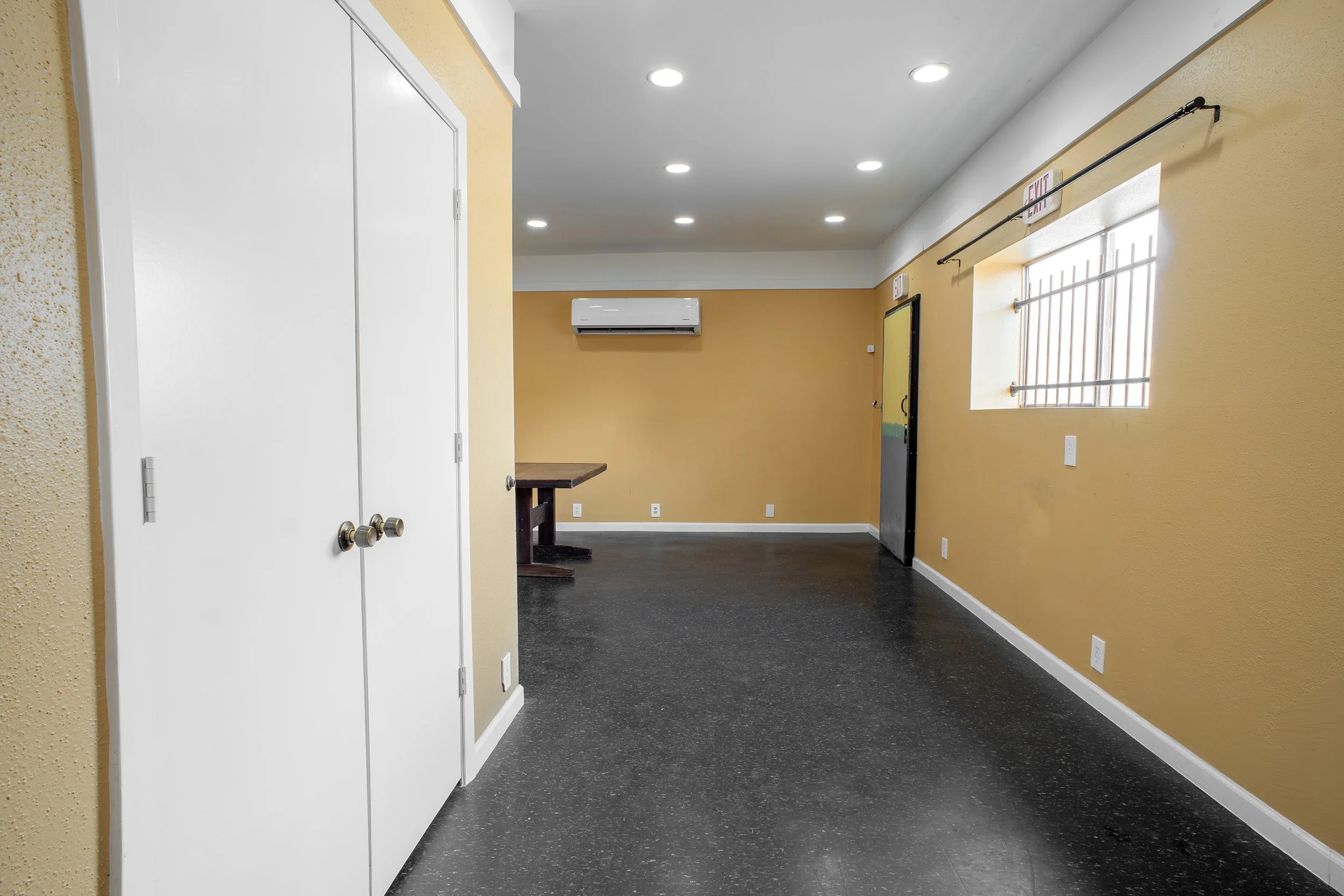 An indoor space with yellow walls, white ceiling with recessed lighting, black flooring, a white air conditioning unit on the back wall, a window with bars, and a doorway with a black frame on the right side.