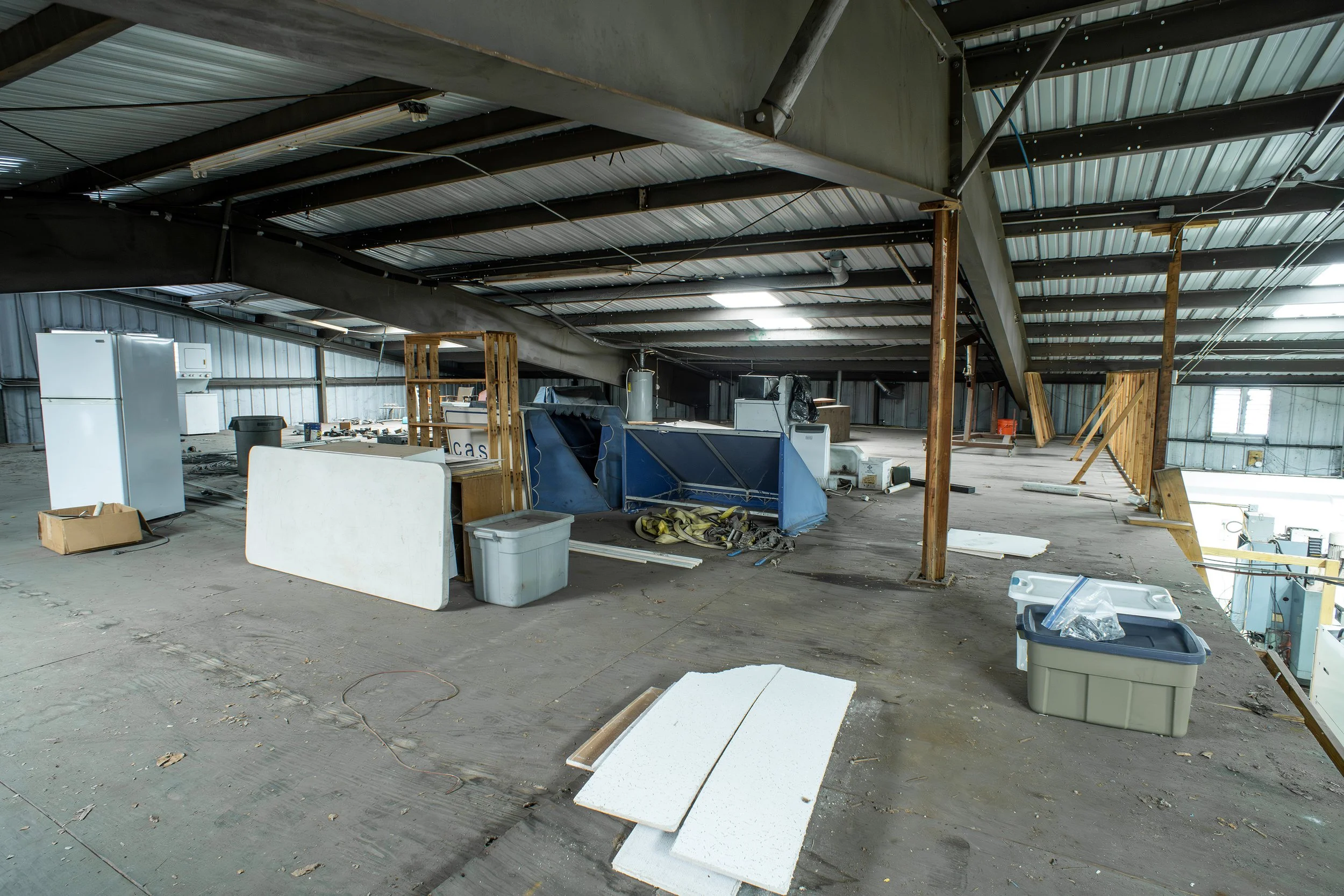Construction or renovation site inside a large industrial building, with installed metal roof, scattered insulation boards, and construction supplies and equipment.