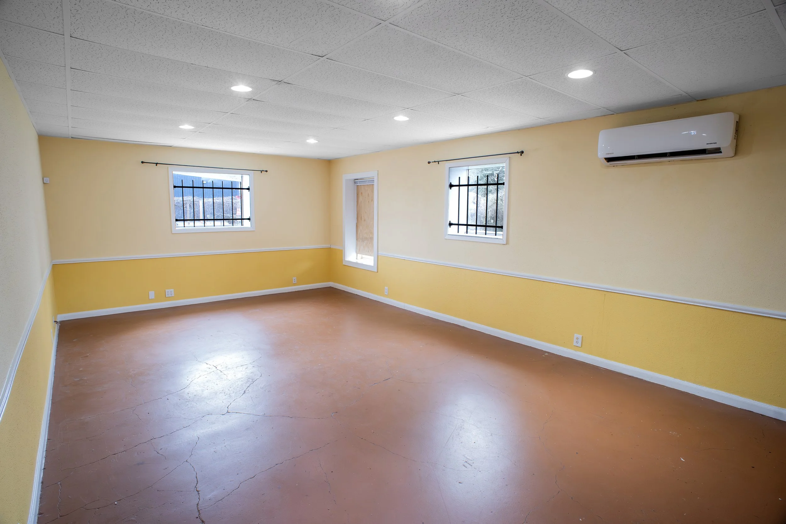 Empty room with yellow walls, two windows with black bars, a door covered with cardboard, a white ceiling with fluorescent lights, and an air conditioning unit on the wall.