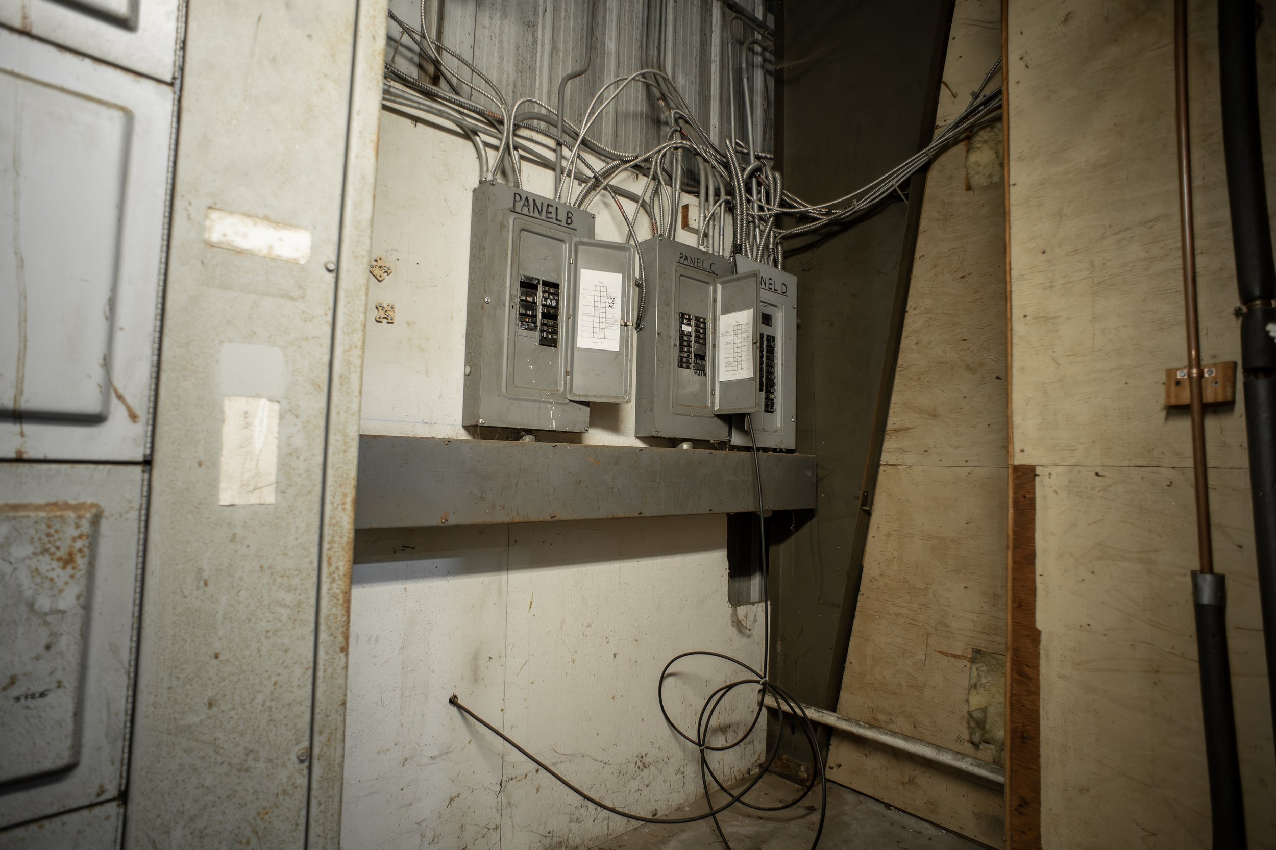 Electrical panels labeled 'Panel B,' 'Panel C,' and 'Panel D' with wires and conduit connected, mounted on a wall in an unfinished or utility room.