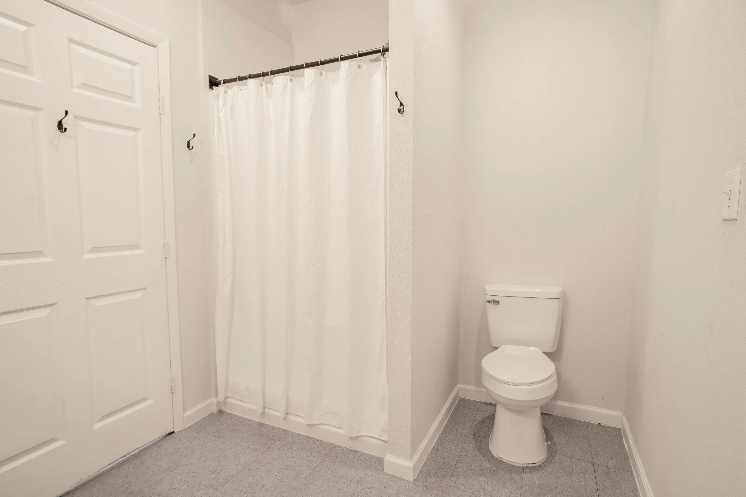 A small bathroom with a toilet, a shower with a white curtain, a white door with hooks, light-colored walls, and gray floor tiles.