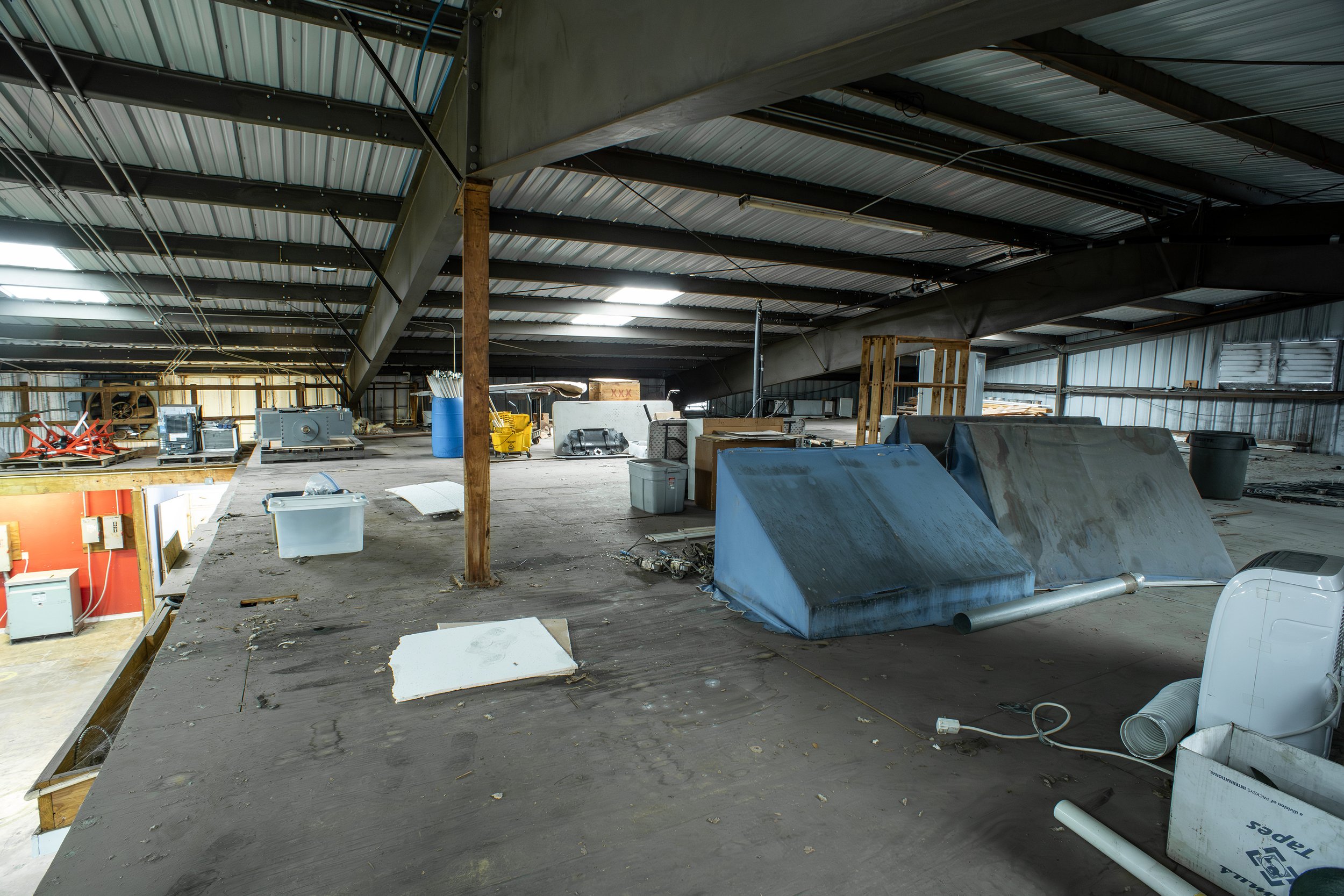 The interior of an unfinished or abandoned building with construction materials, debris, and equipment scattered around. The ceiling has metal panels supported by steel beams, and part of the floor is unfinished with exposed framing and insulation.