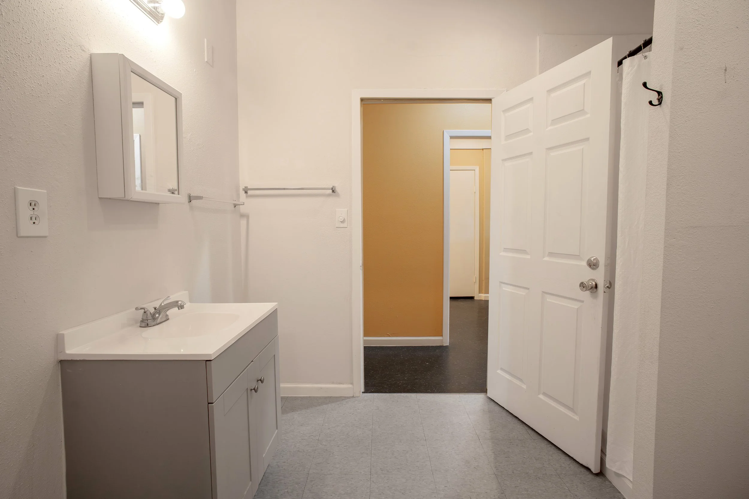 An interior view of a bathroom with a white sink vanity, mirror, towel bar, and open door leading to a hallway with beige walls.