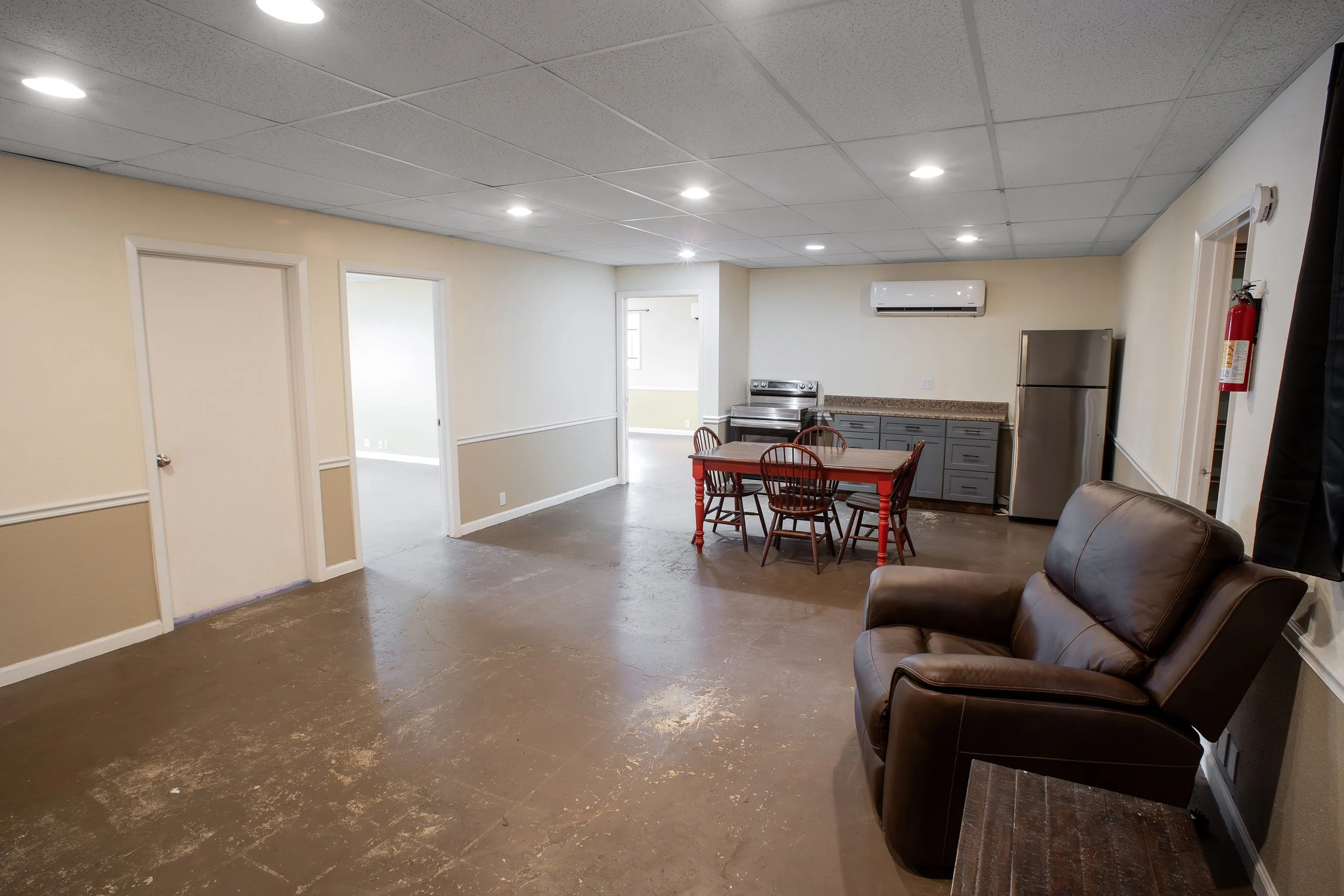 Interior of a room with beige walls, a tile suspended ceiling with recessed lighting, a small kitchen area with gray cabinets, a stainless steel fridge, and a countertop, a wooden table with four chairs, a brown leather sofa, and a window on the far 