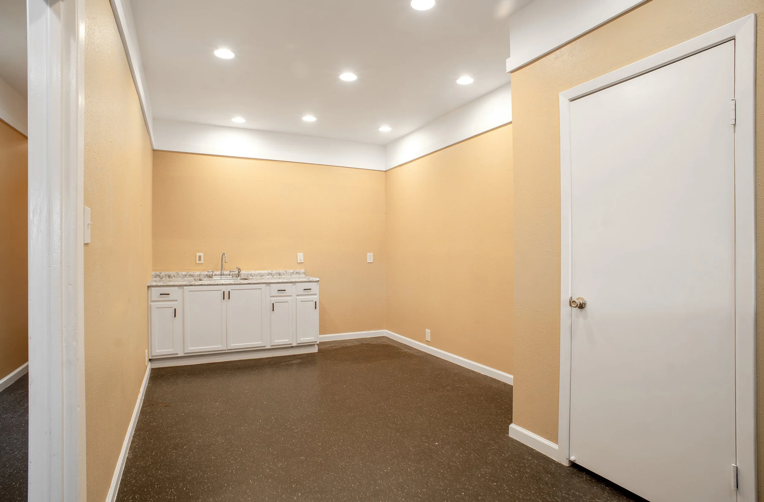 Empty room with beige walls, white ceiling with recessed lighting, dark speckled carpet, a small cabinet with a sink, and a white door.