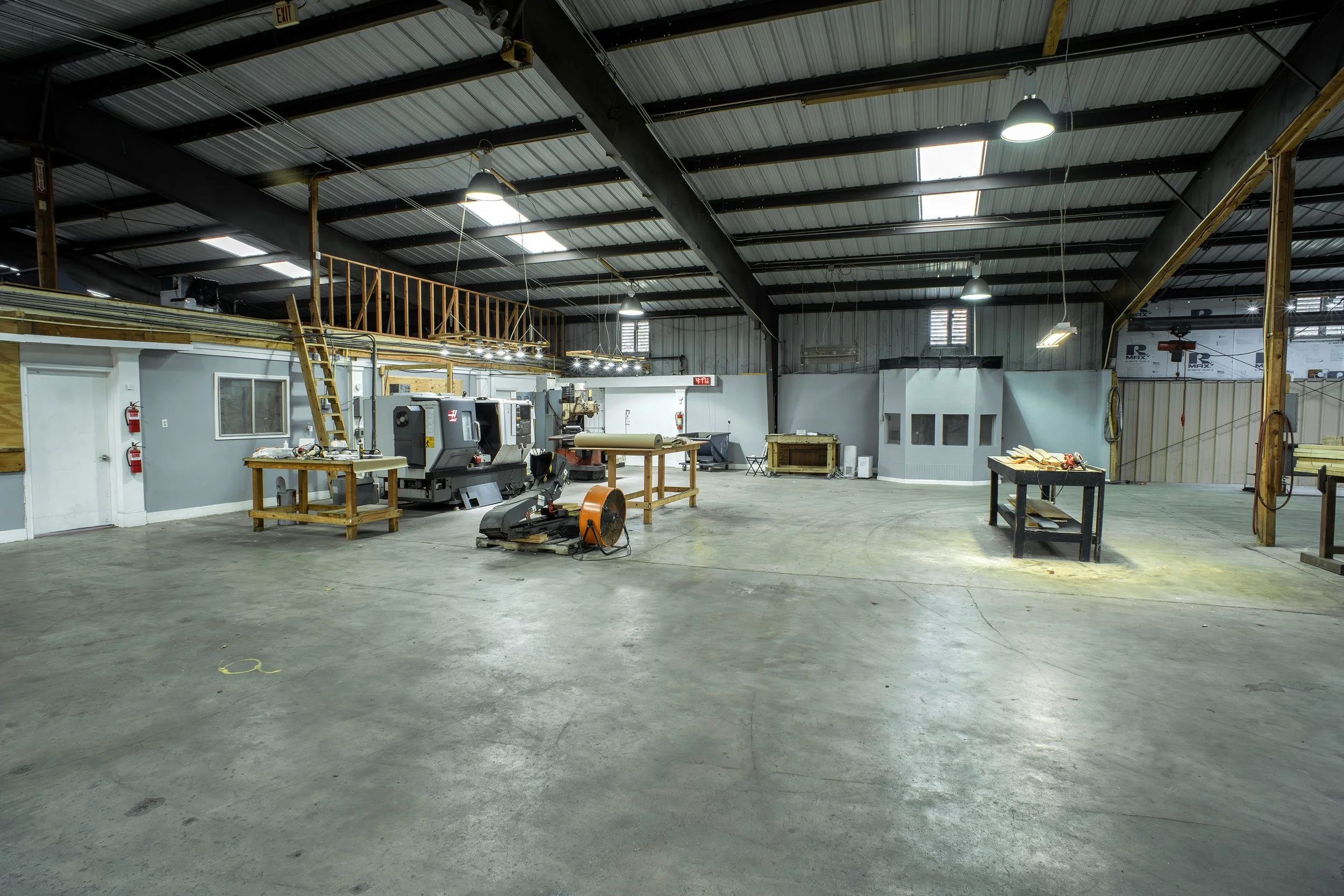 An empty industrial workshop with metal ceiling, skylights, fluorescent lights, and woodworking tools and machinery scattered across a concrete floor.