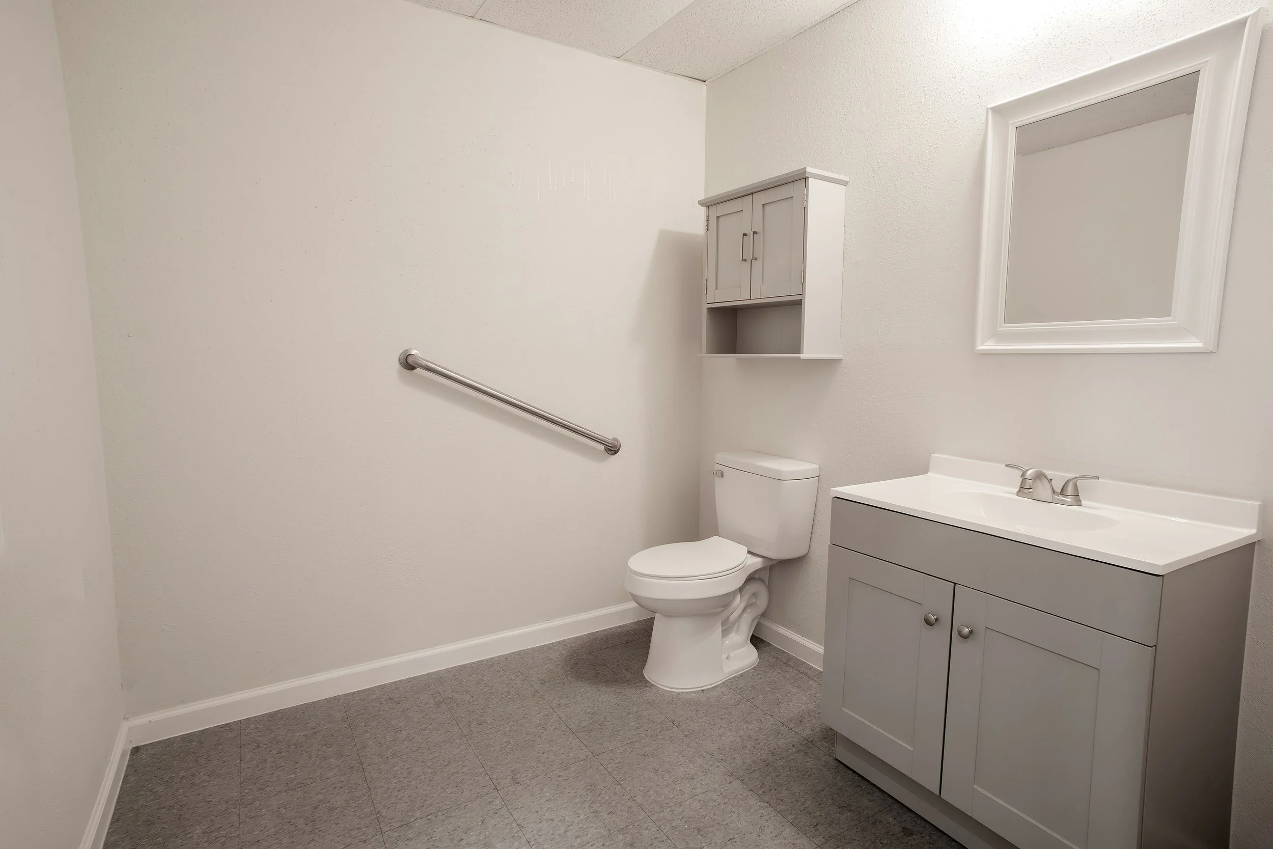 A clean and minimalistic bathroom with white walls, a toilet, a gray vanity with a sink, a large mirror framed in white, a wall-mounted cabinet, and a handrail along one wall.