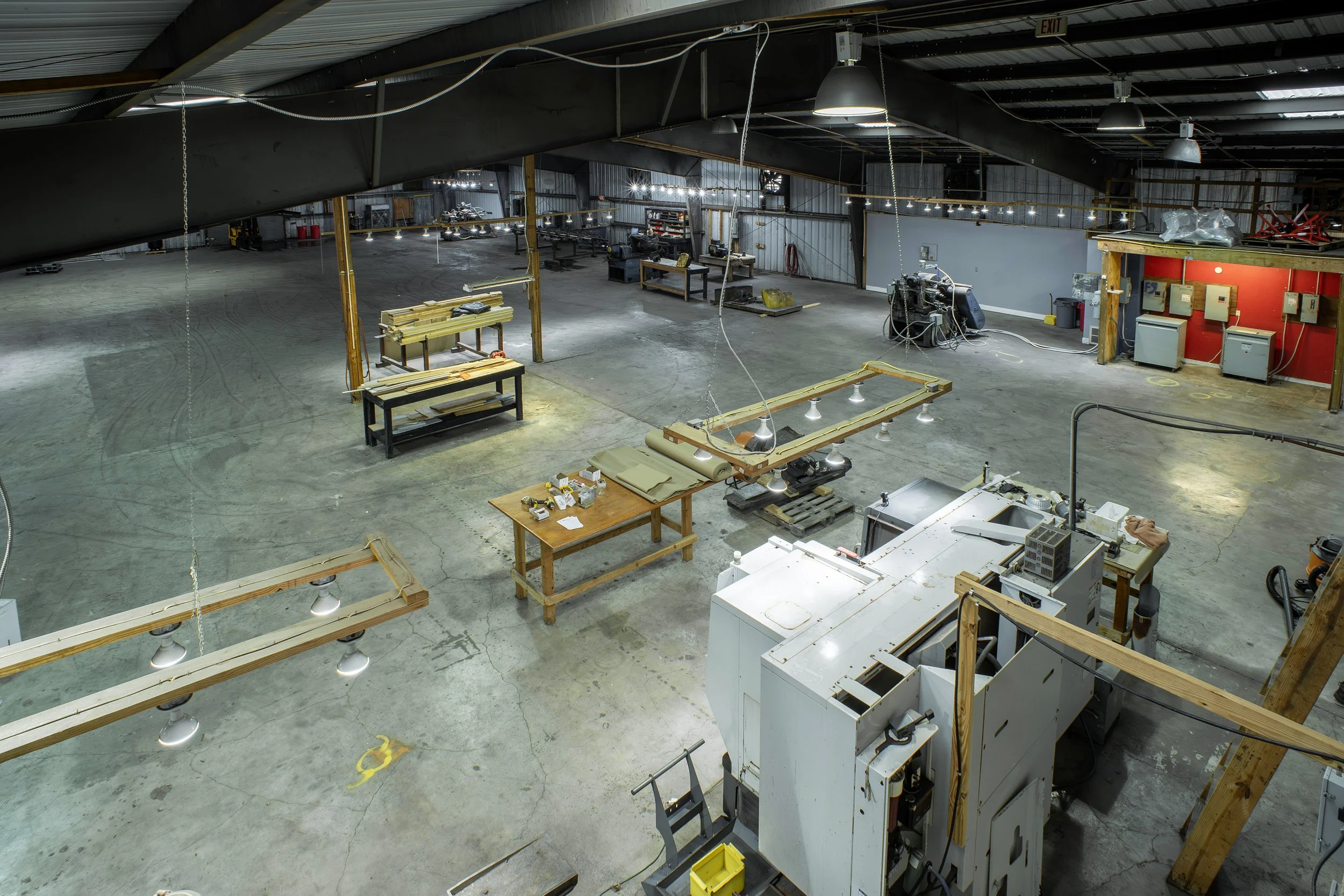 Empty warehouse or workshop with workbenches, tools, equipment, and scattered materials. Overhead lighting and exposed ceiling structure.