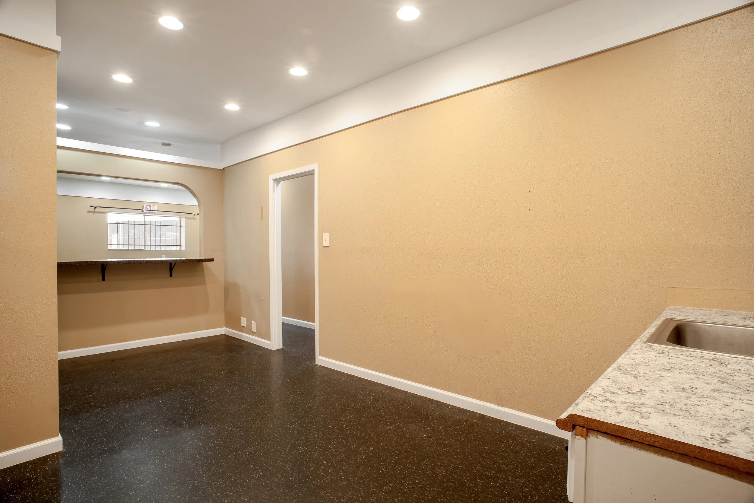 An interior view of a modern kitchen and living area with beige walls, white ceiling lights, a beige and white granite countertop, a small sink, a door, a window with bars, and a counter with a granite top.