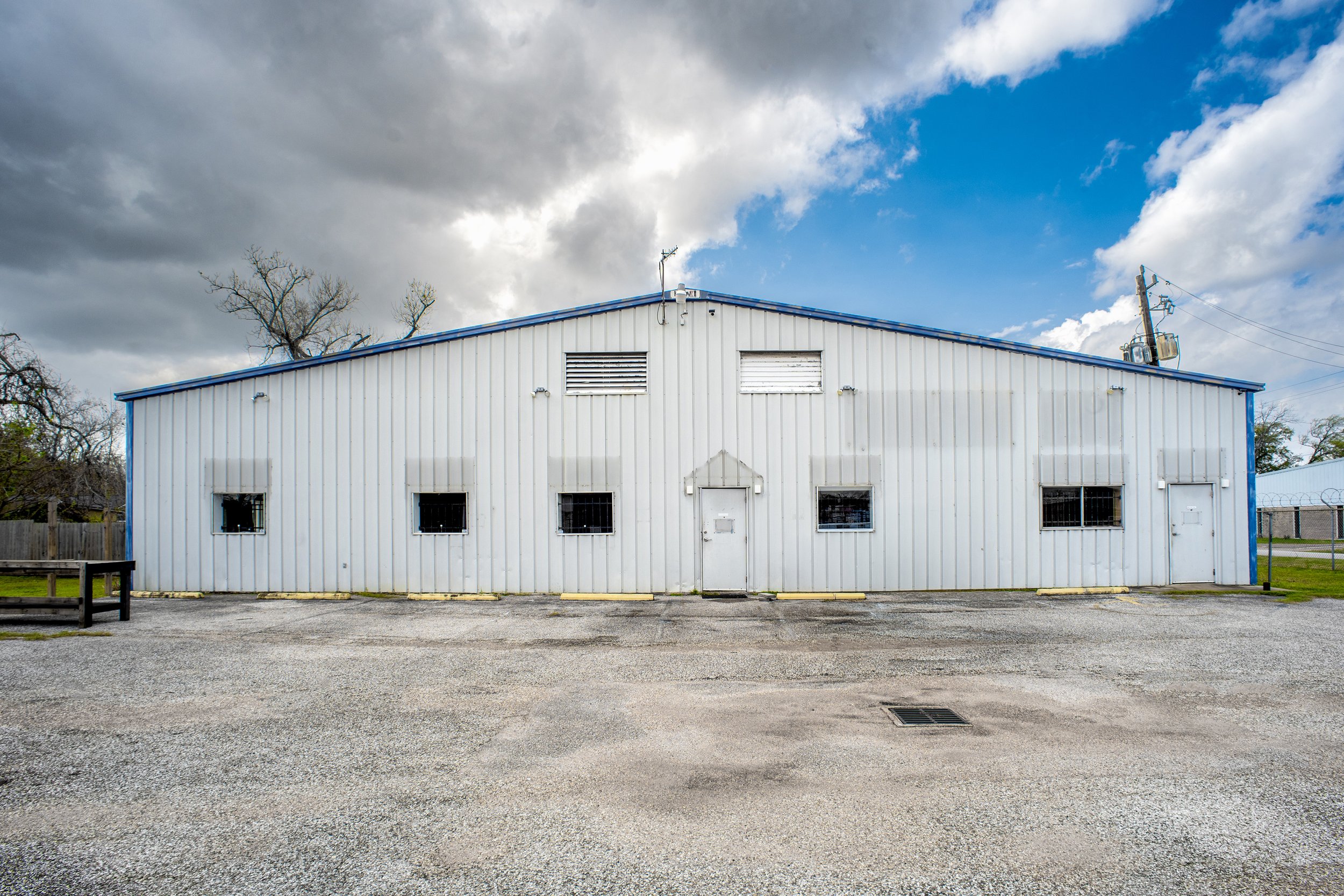 White industrial building with a blue roof, several small windows, and vents, set against a cloudy sky with some blue patches, surrounded by a parking lot and a few trees.