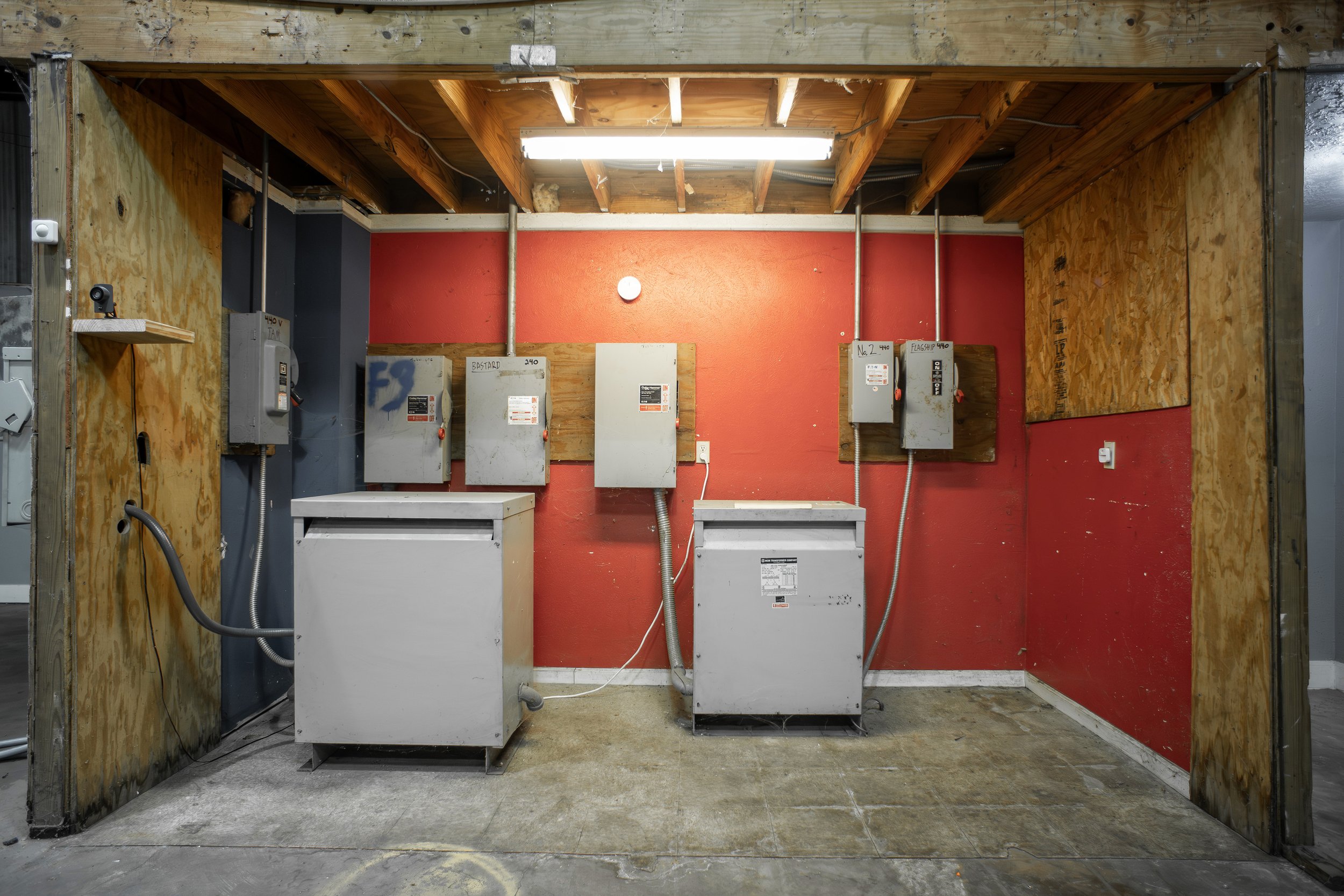 Construction site with electrical panels and exposed wooden ceiling, partially enclosed with wood and drywall.