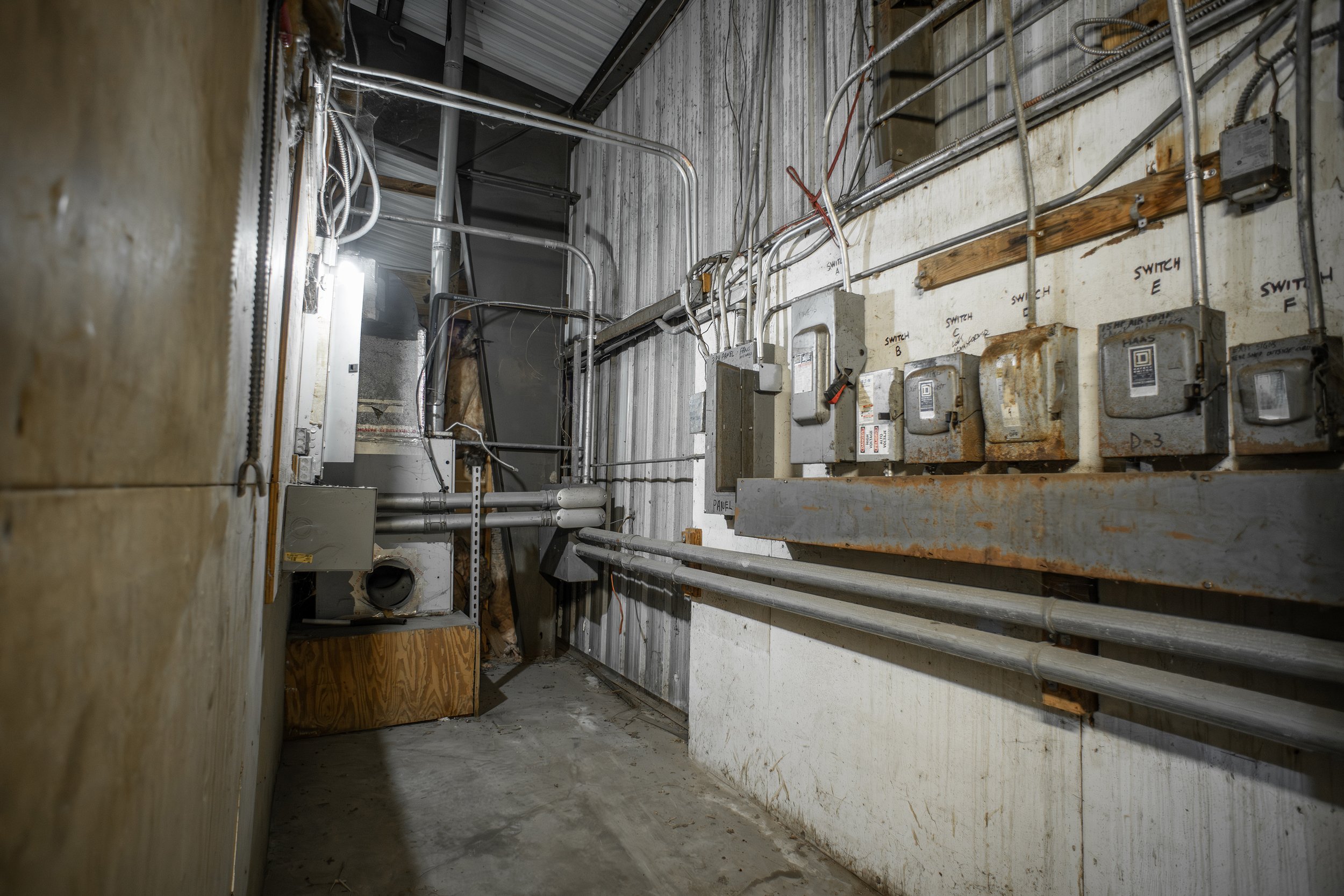 Construction or utility room with exposed electrical wiring and panels on a white wall, various tubes and pipes, some labeled, and unfinished surfaces.