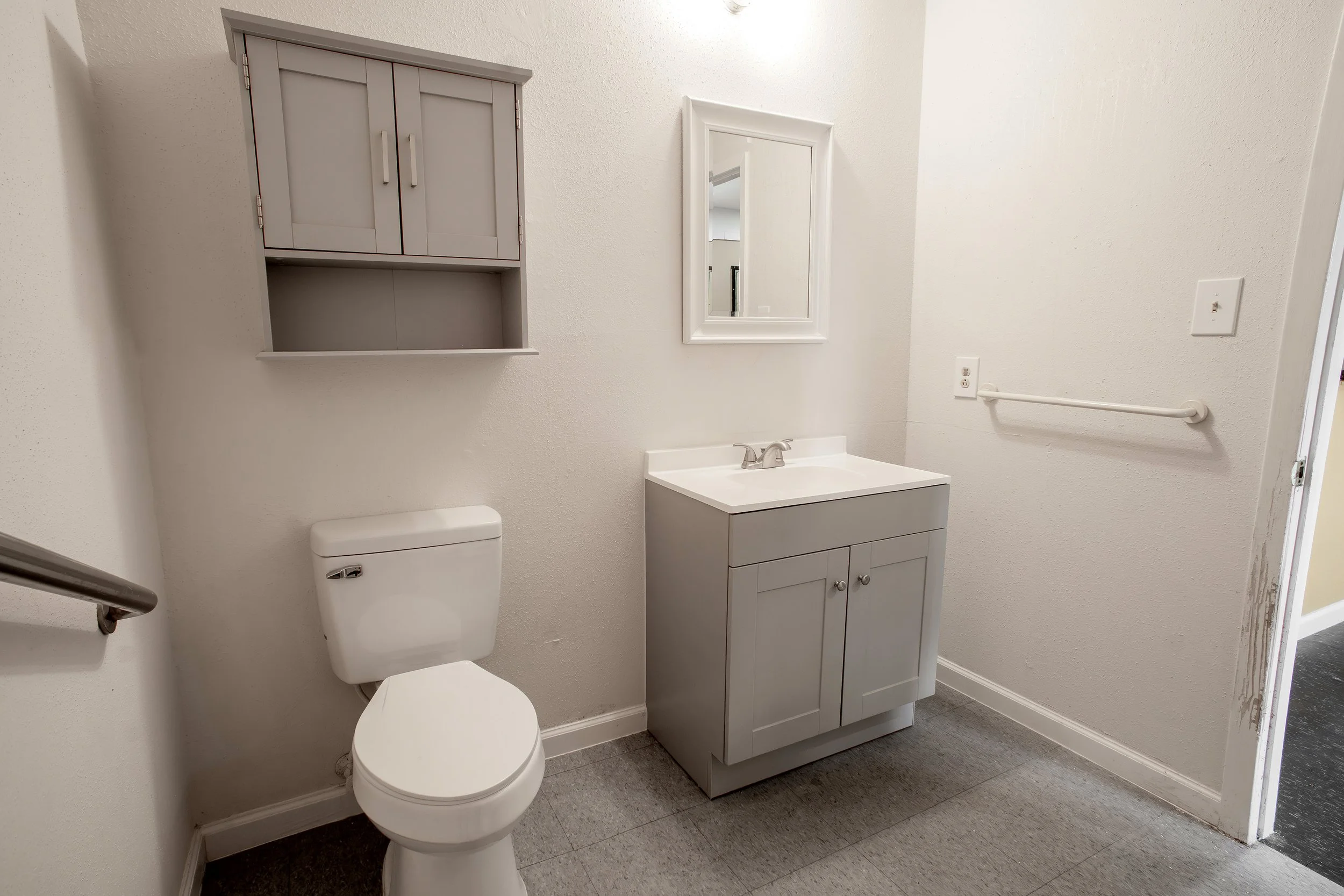 A small bathroom with a white toilet, a gray vanity with a sink, a mirror, an over-the-toilet cabinet, a white wall, and a textured white wall.