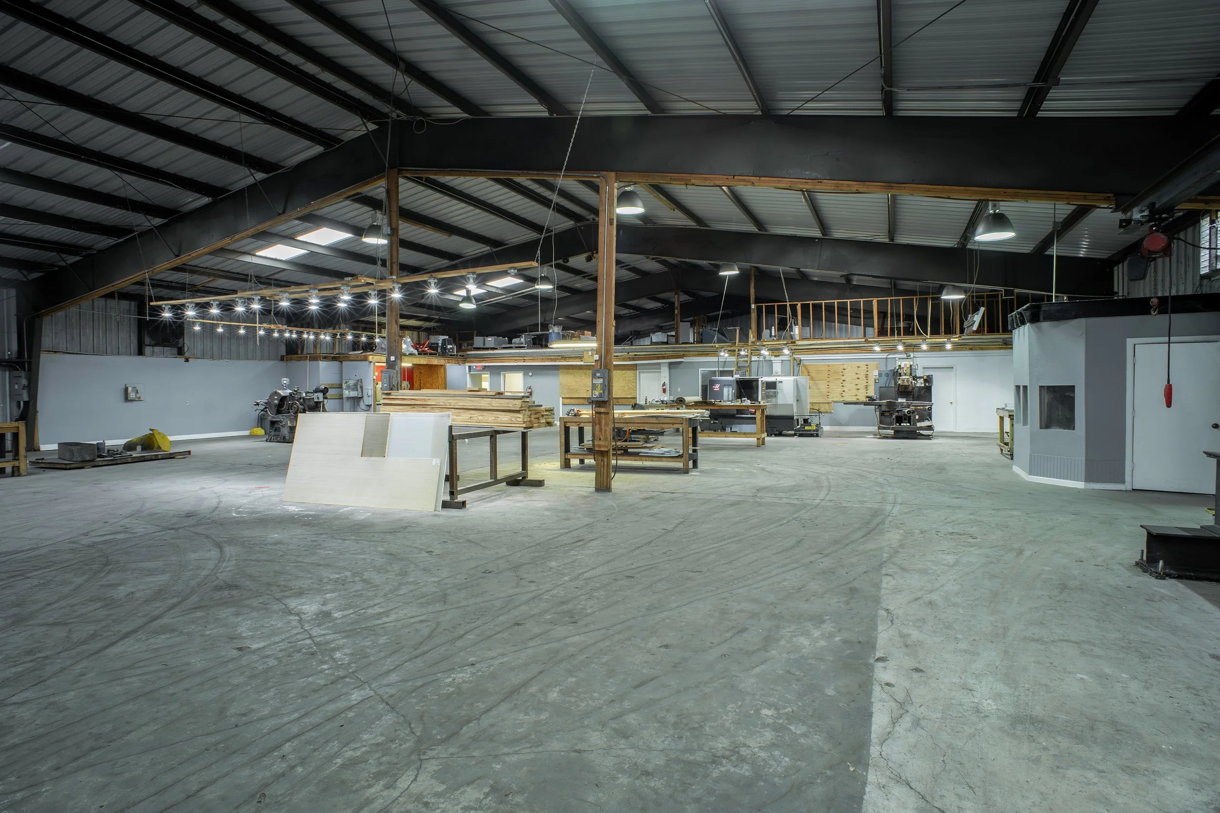 Interior of a large industrial workshop or warehouse with metal roofing, wooden beams, workbenches, and industrial machinery, illuminated by ceiling lights.