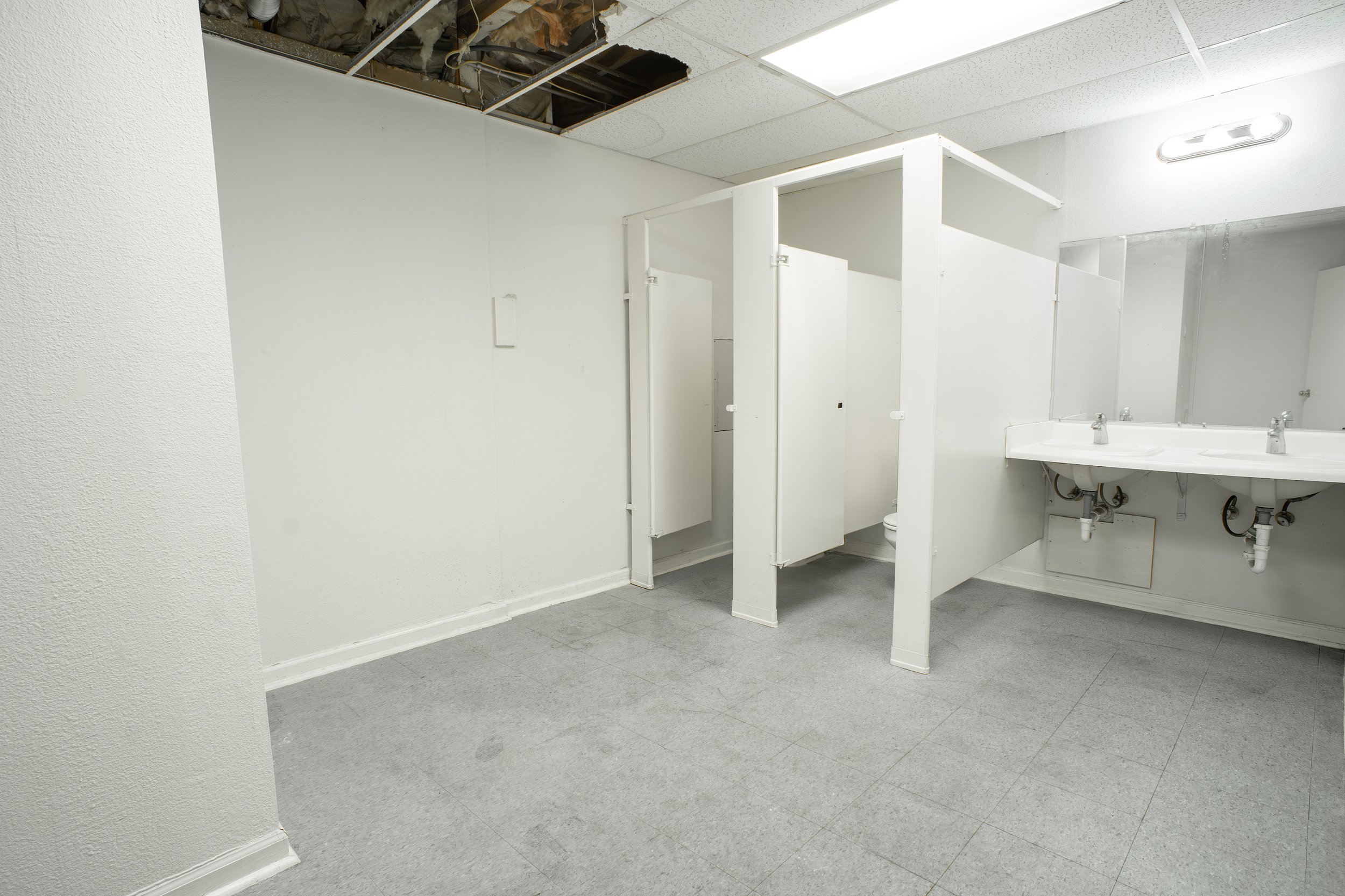 Empty men's or women's public bathroom with white walls, gray flooring, a double sink with mirrors, and several stalls, one of which is under construction or renovation with missing ceiling tiles.